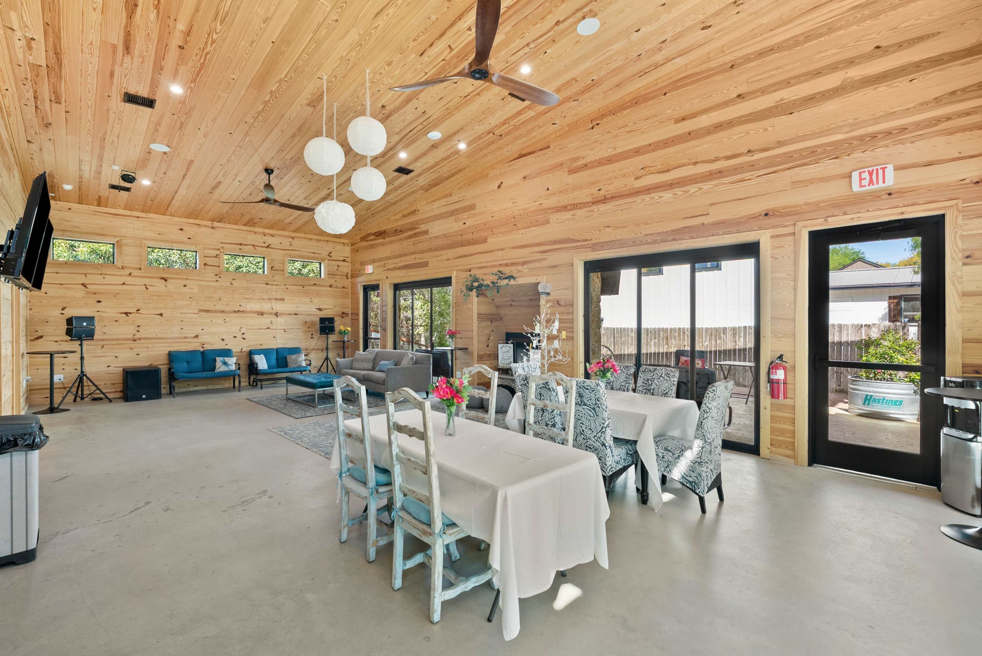 The image shows a spacious wooden interior with a dining table set for guests, lounge seating, and large windows providing natural light.