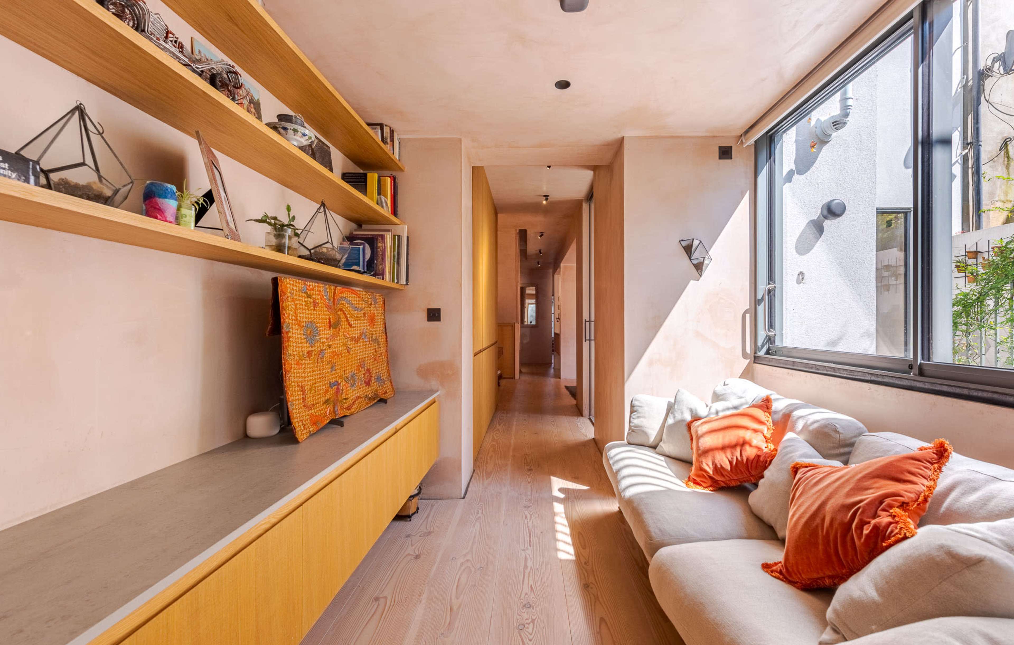 The image shows a narrow, well-lit hallway featuring a living area with a light-colored sofa adorned with orange pillows and a wooden shelf displaying decorative items.