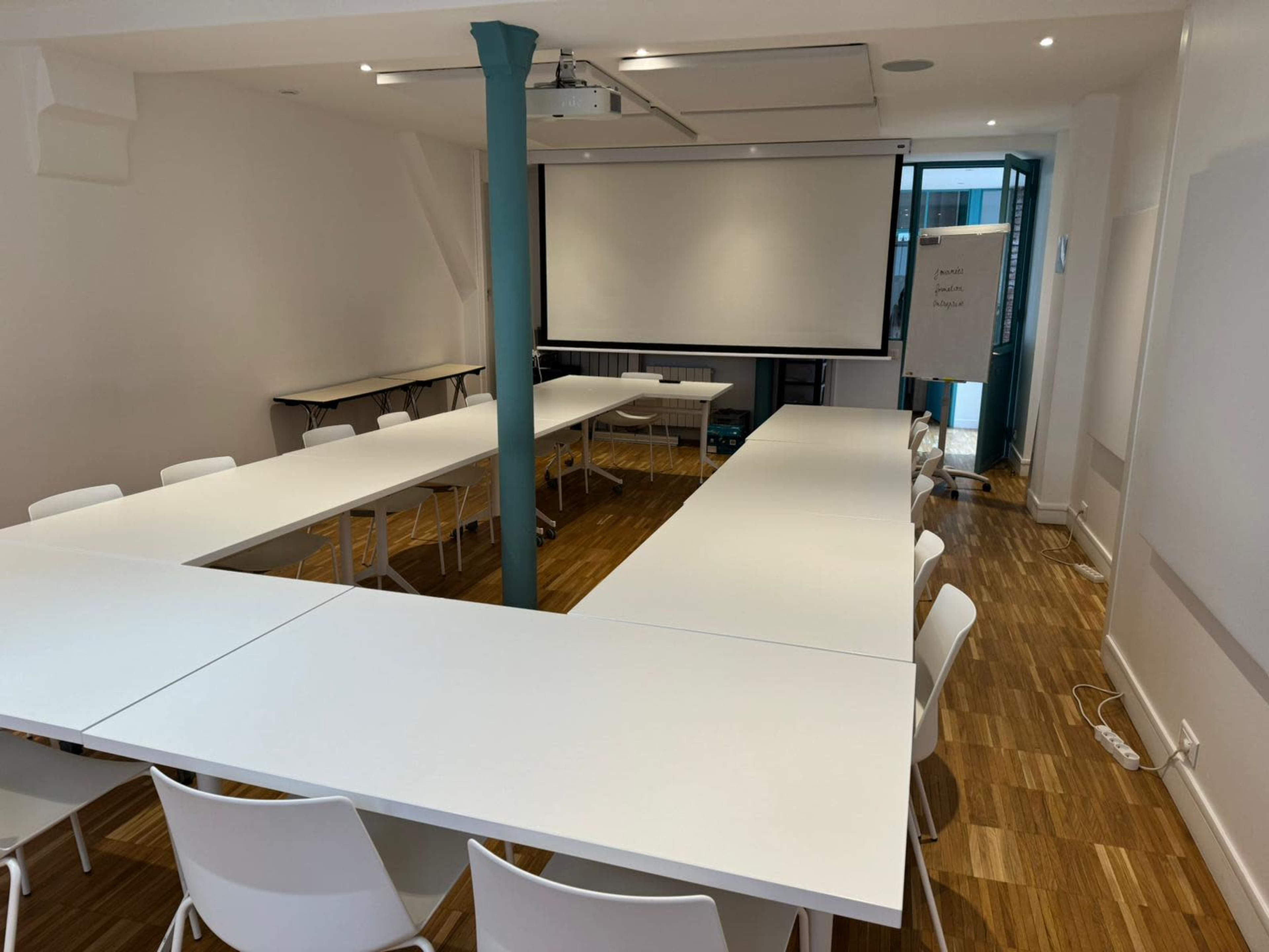 A meeting room with white tables arranged in a U-shape and several white chairs, featuring a projector screen and large windows.
