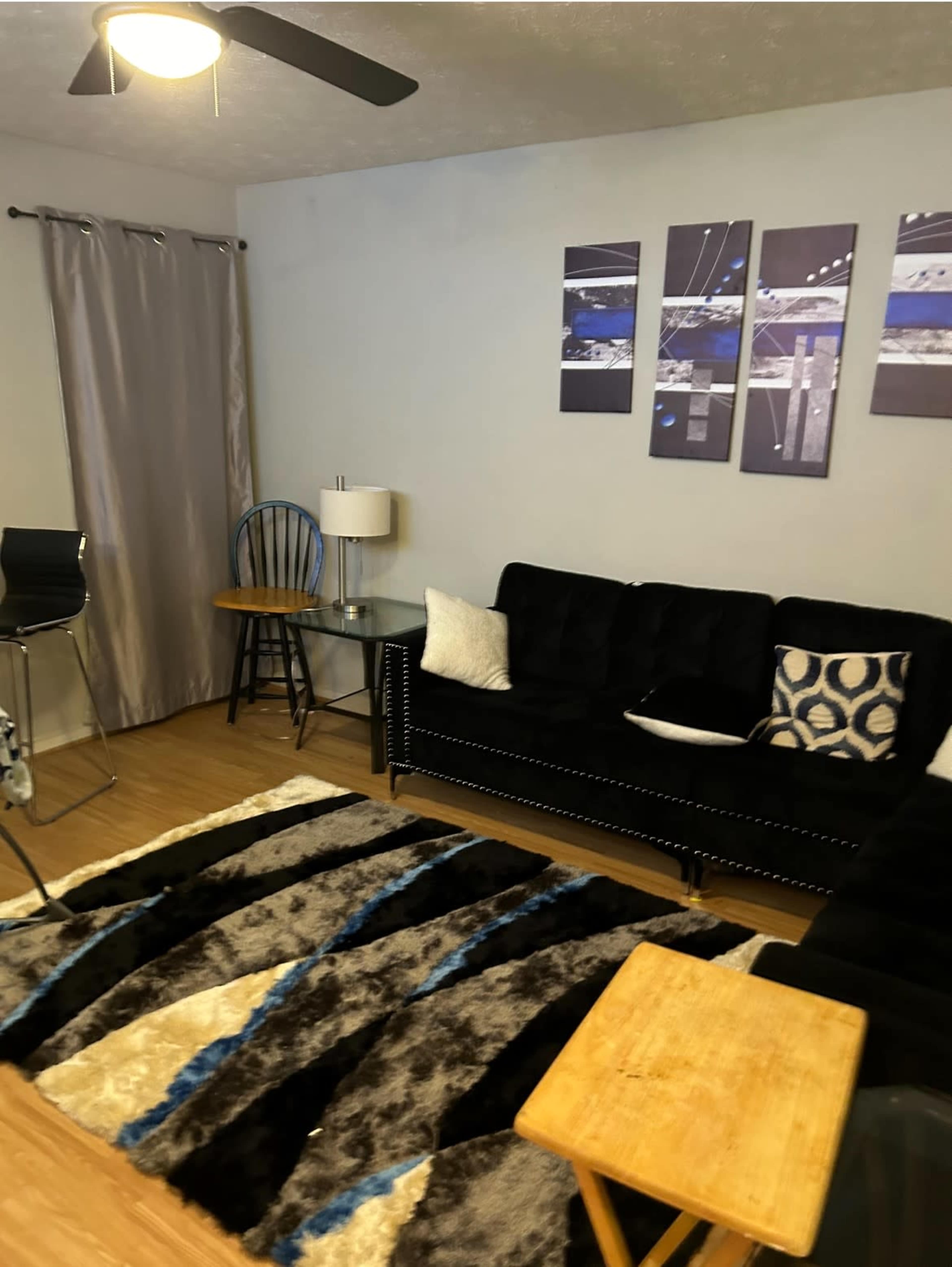 The image shows a living room featuring a black sofa, a glass side table, a lamp, a blue and black rug, and wall art, with a curtain covering a window.