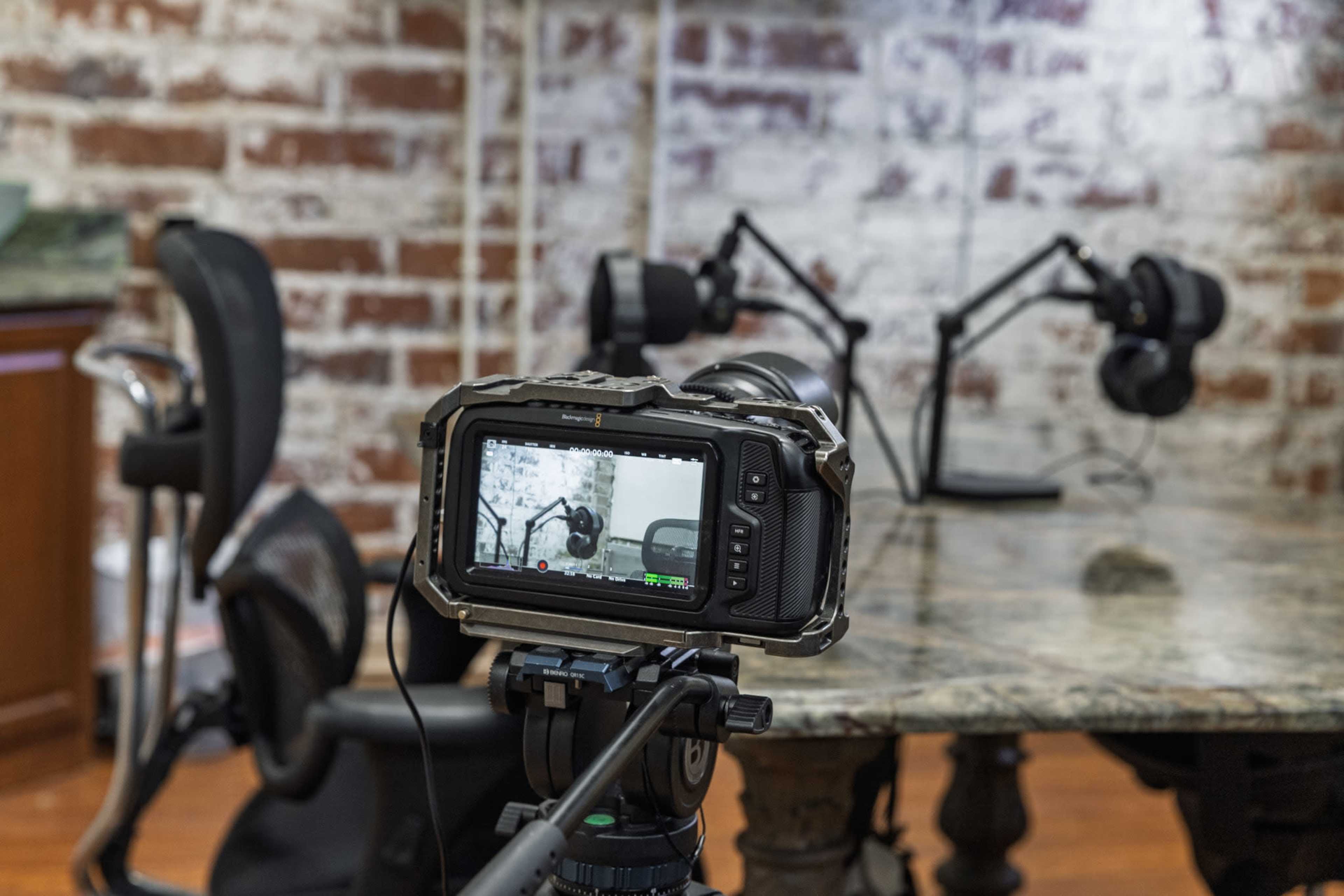 A camera on a tripod captures a tabletop with two microphones and headphones in a room with exposed brick walls.