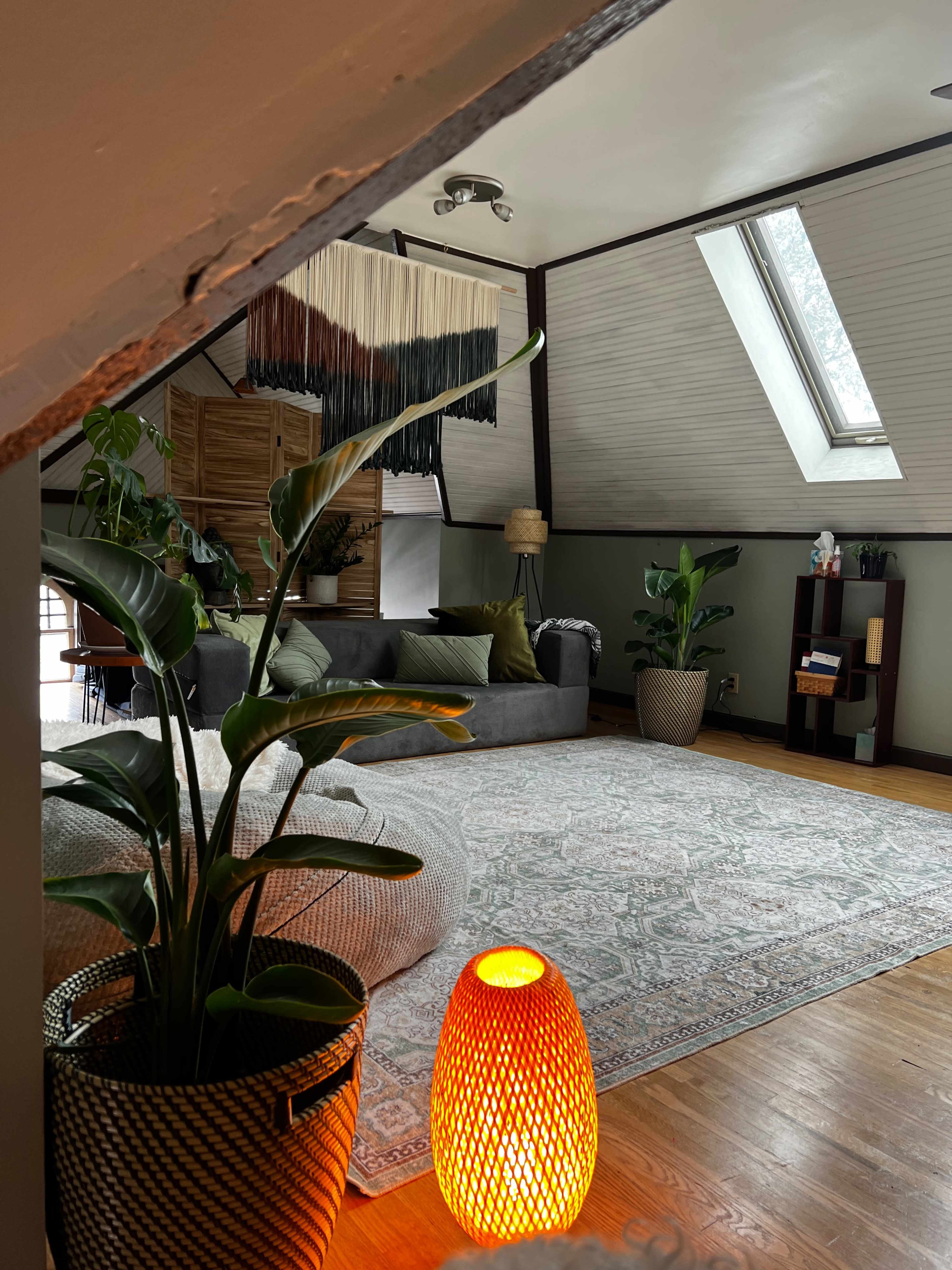 A cozy attic living room features a gray sofa, a textured area rug, potted plants, and a unique amber lamp.