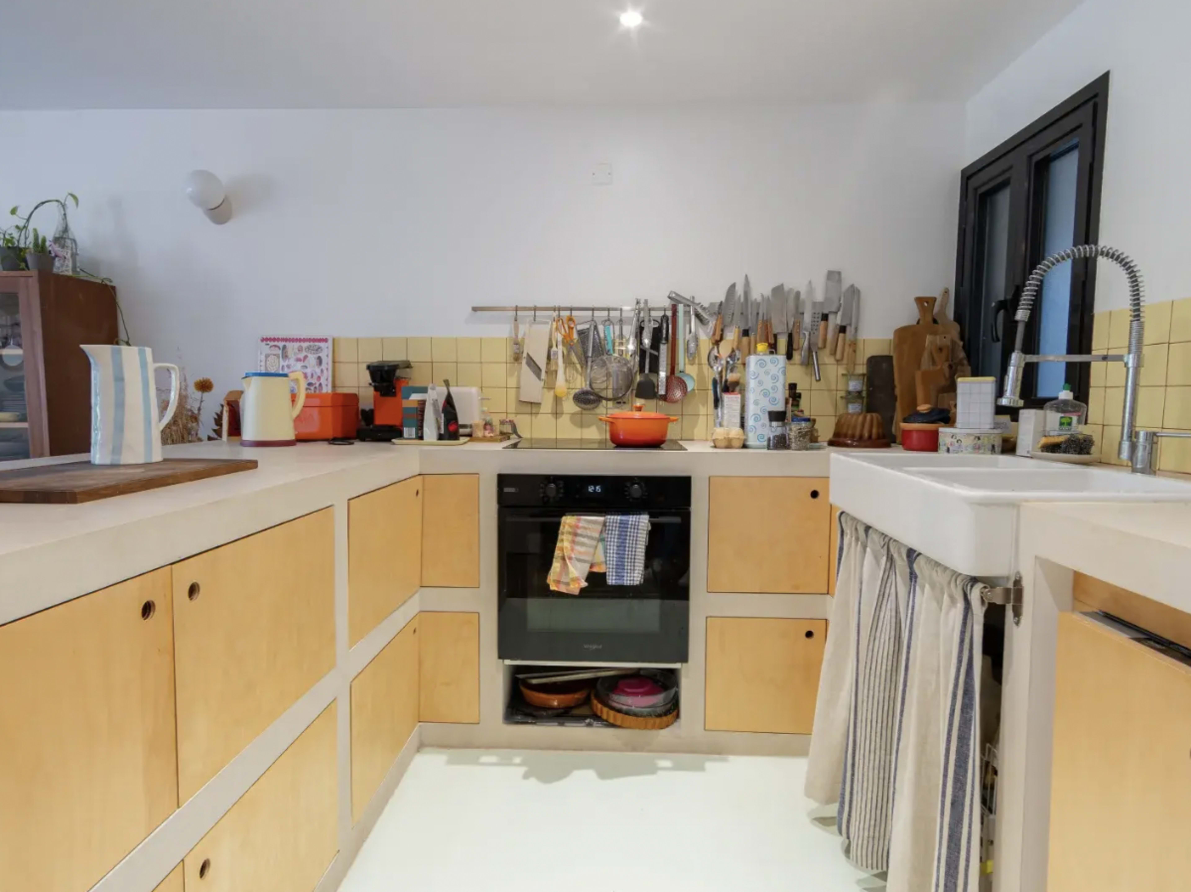 The image shows a modern kitchen with light wood cabinets, a black oven, and a variety of cooking utensils displayed on the wall.