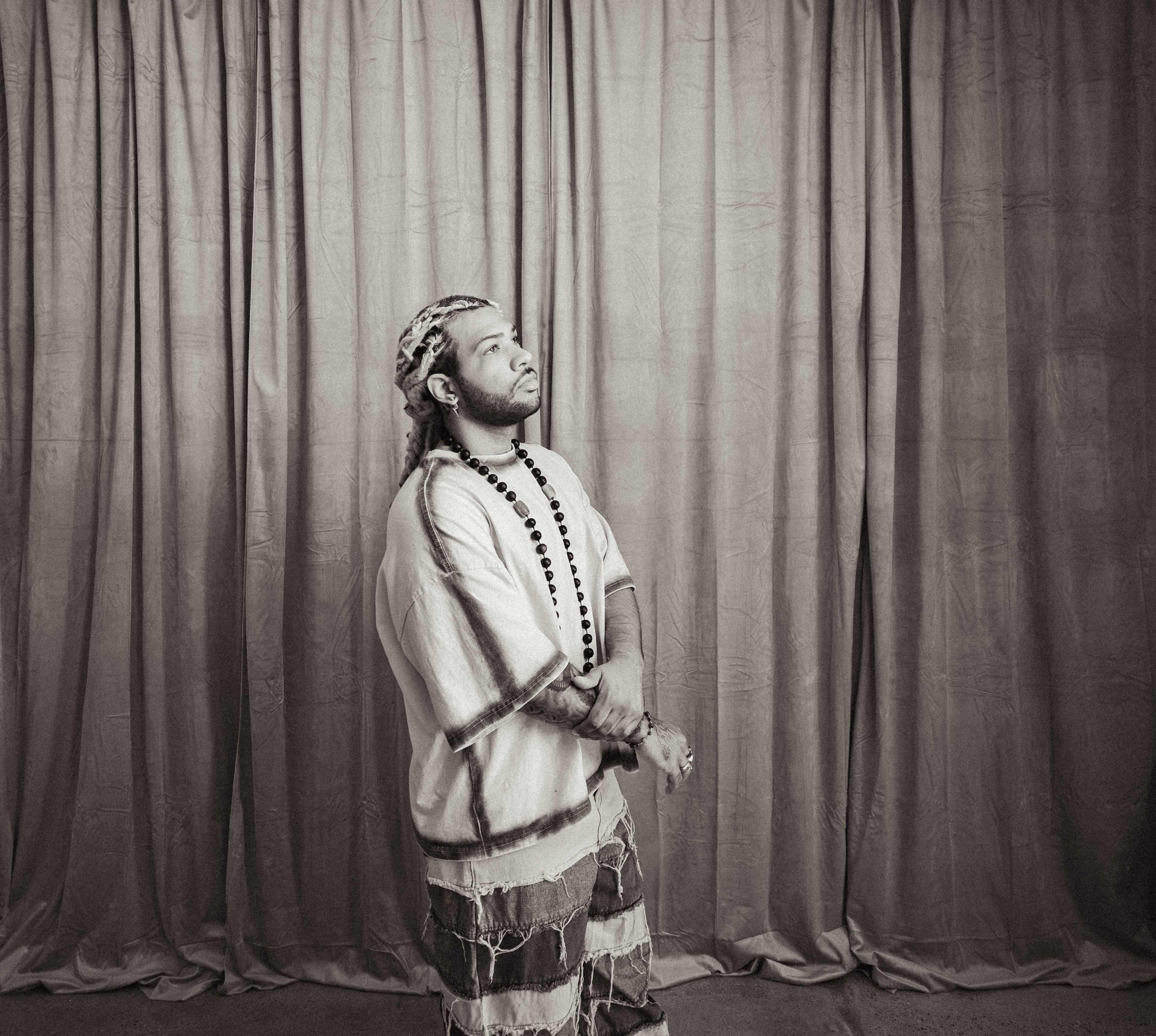 A man dressed in traditional attire stands in front of a textured curtain backdrop.