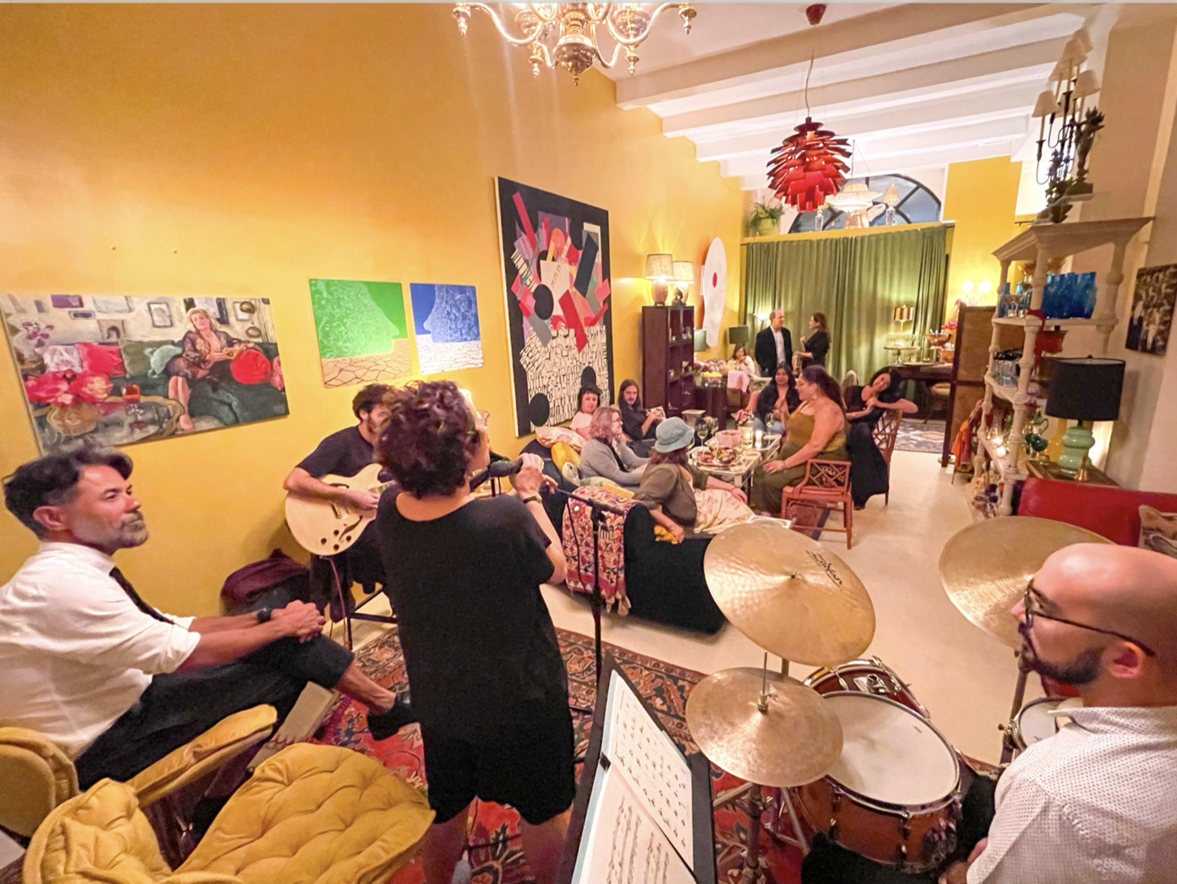 A group of musicians and guests gather in a colorful living room, with instruments set up for a jam session amid vibrant artwork and decor.