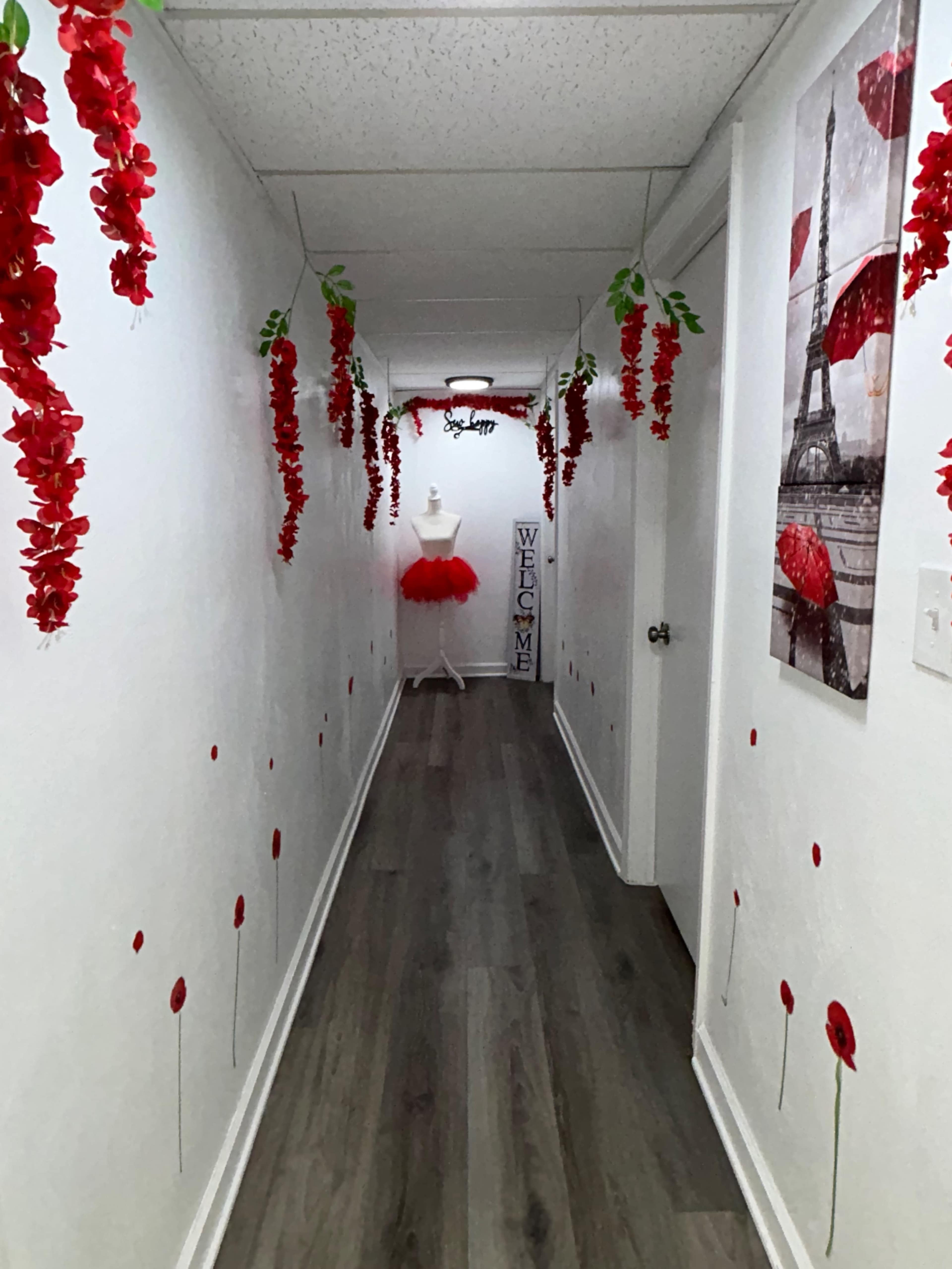 A narrow hallway is decorated with red flowers hanging from the ceiling and images of poppies on the walls, leading to a mannequin wearing a tutu and a "Welcome" sign at the end.