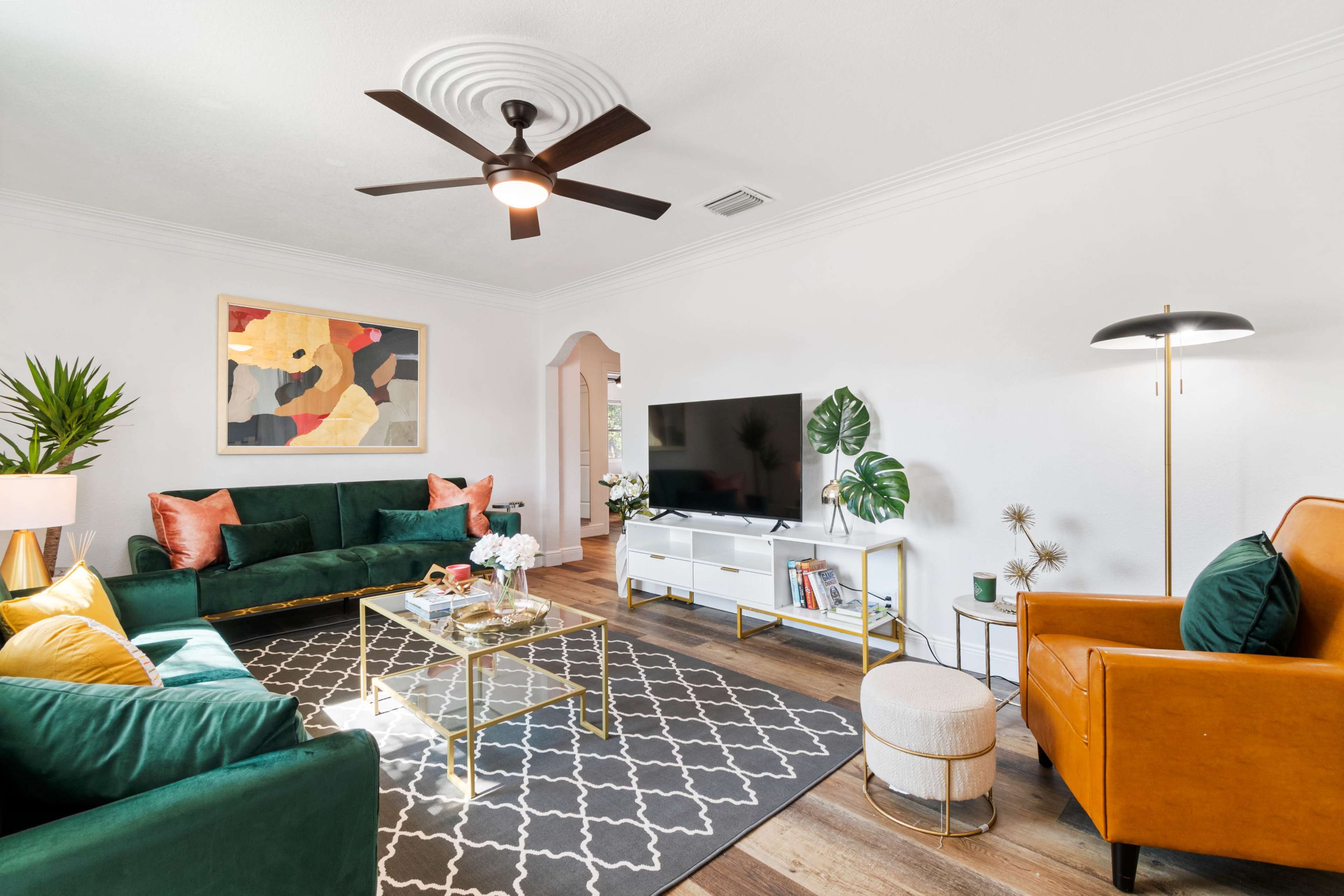 The living room features a green sectional sofa, a modern coffee table, a television on a white stand, and a combination of decorative plants and lighting.