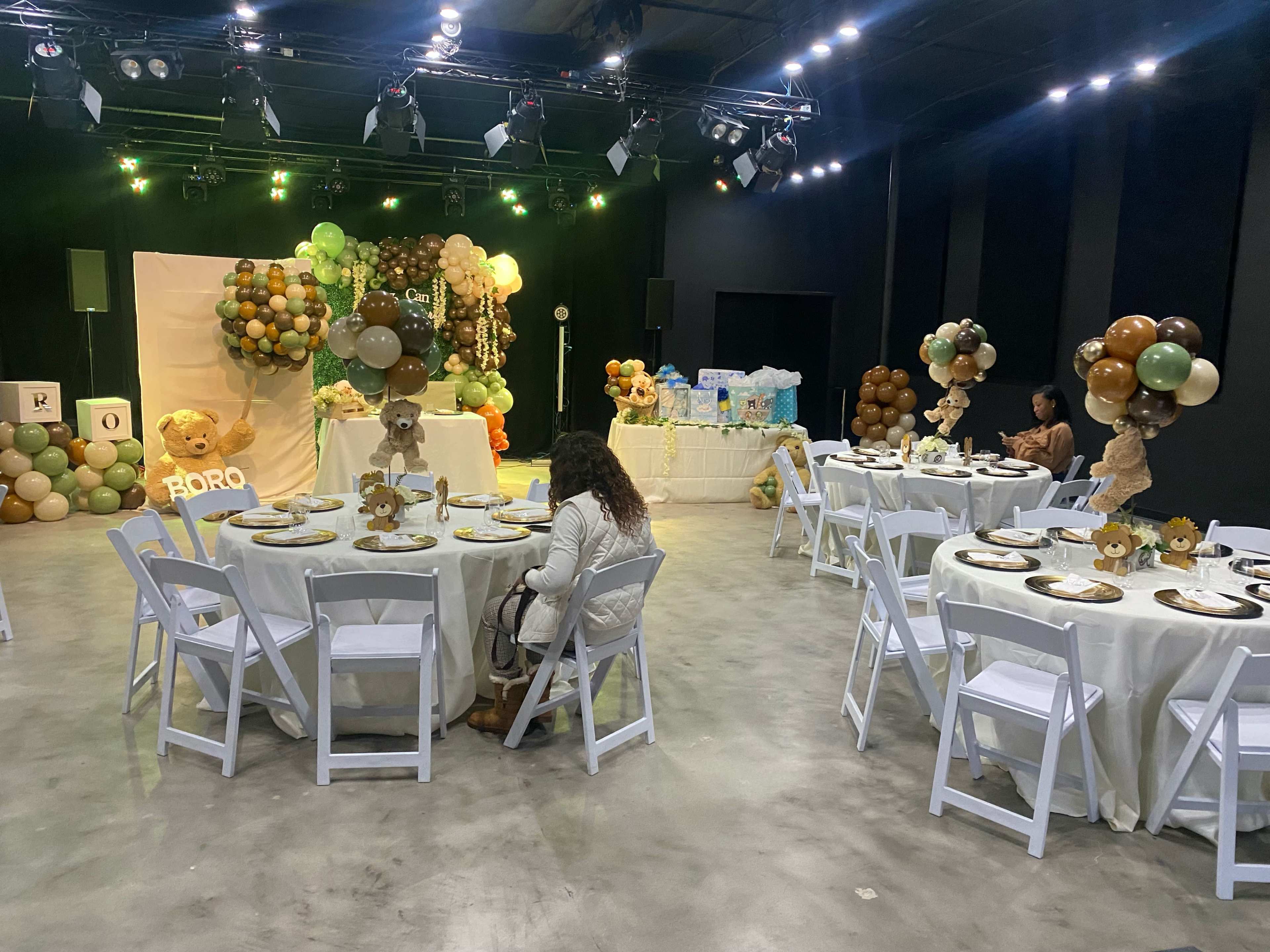 A decorated event space features round tables set with plates and centerpieces, while a display of balloons and gifts is arranged at the back.