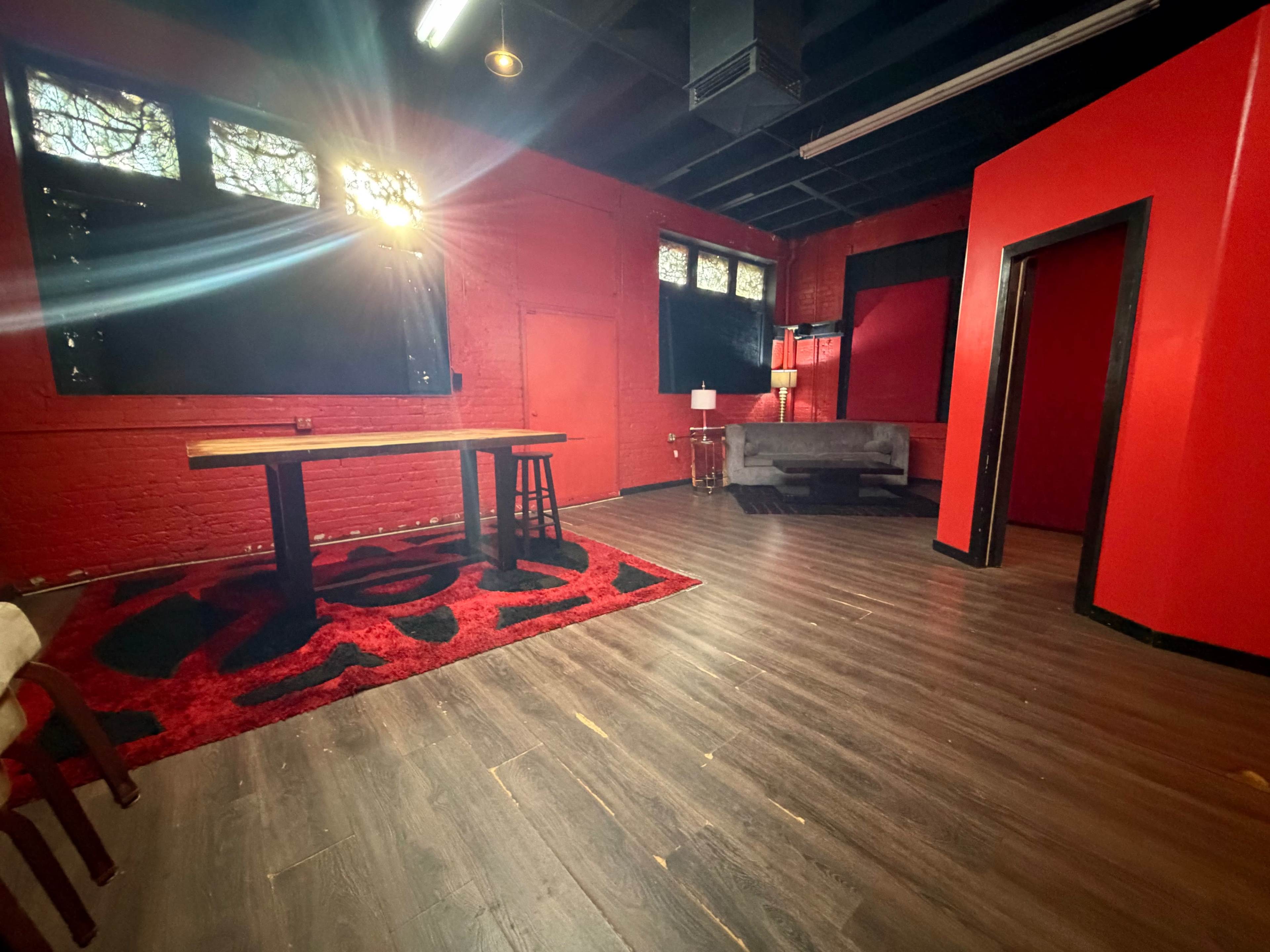 The room features red walls, a wooden table with a stool, a gray couch, and a large rug on the floor.