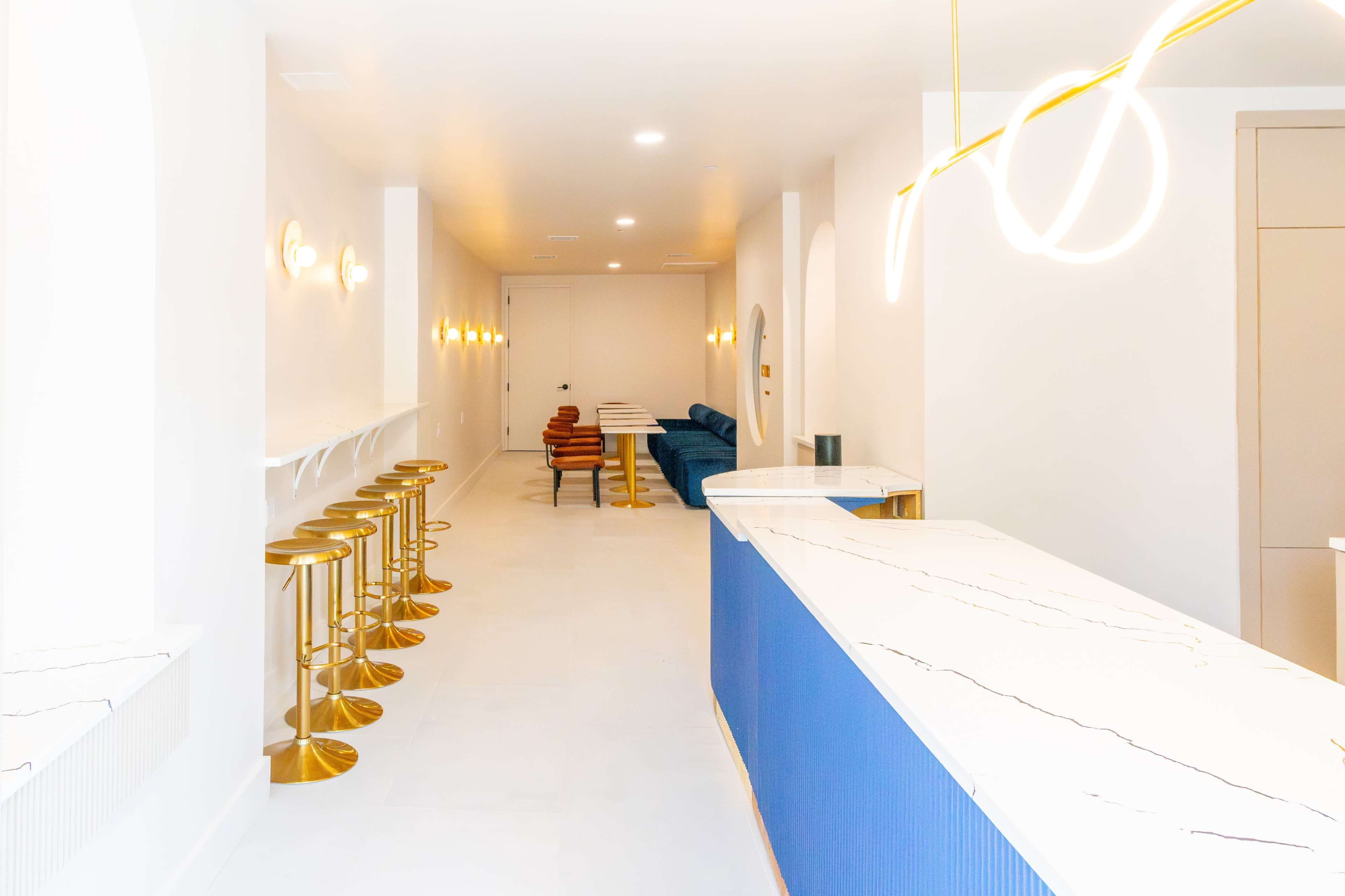The image shows a modern, brightly lit interior of a café or lounge, featuring a long white marble bar with blue accents, several gold stools, and a row of light fixtures along the walls.