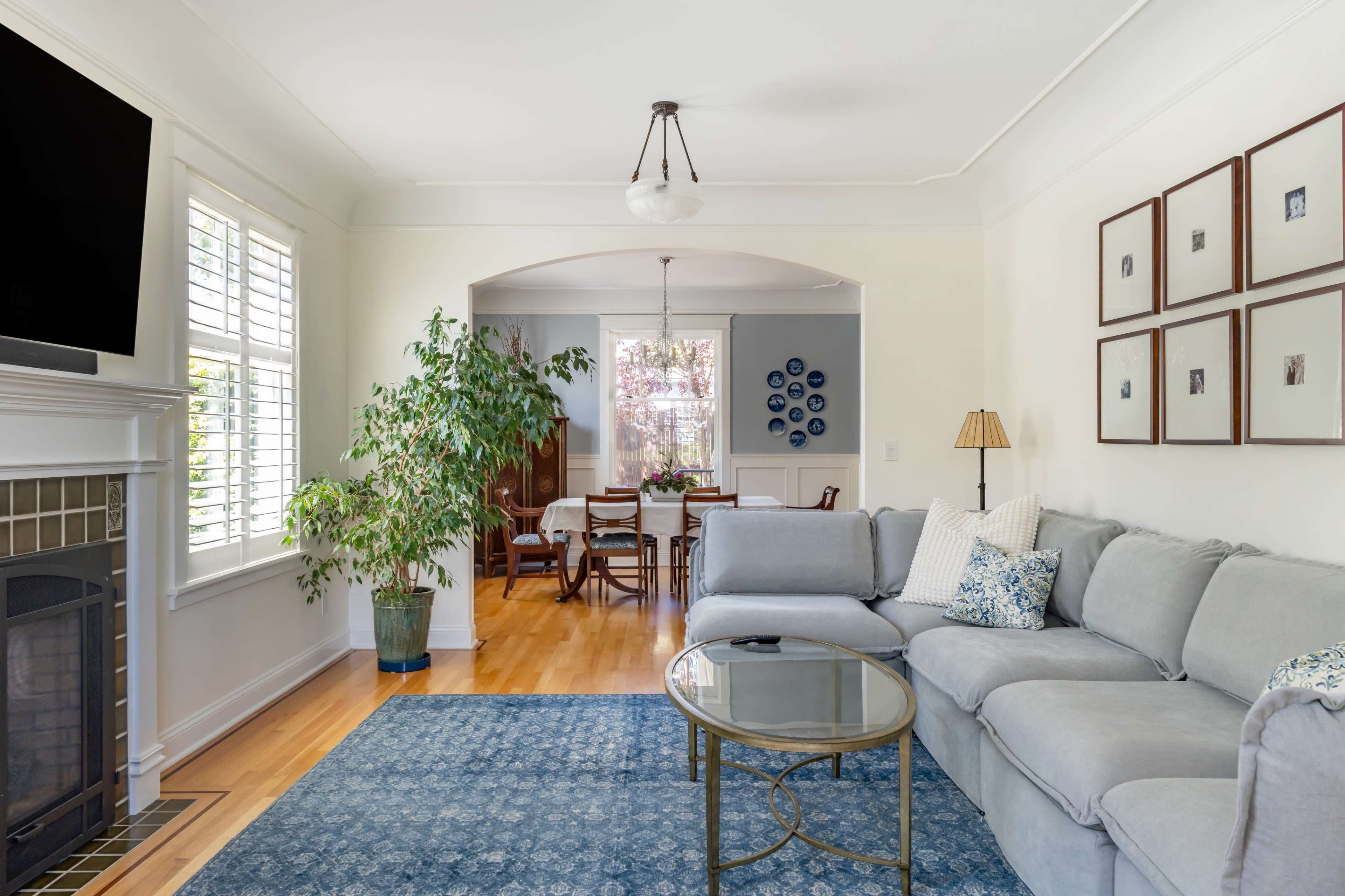A modern living room features a gray sectional sofa, a round glass coffee table, and a fireplace, with a dining area visible in the background.
