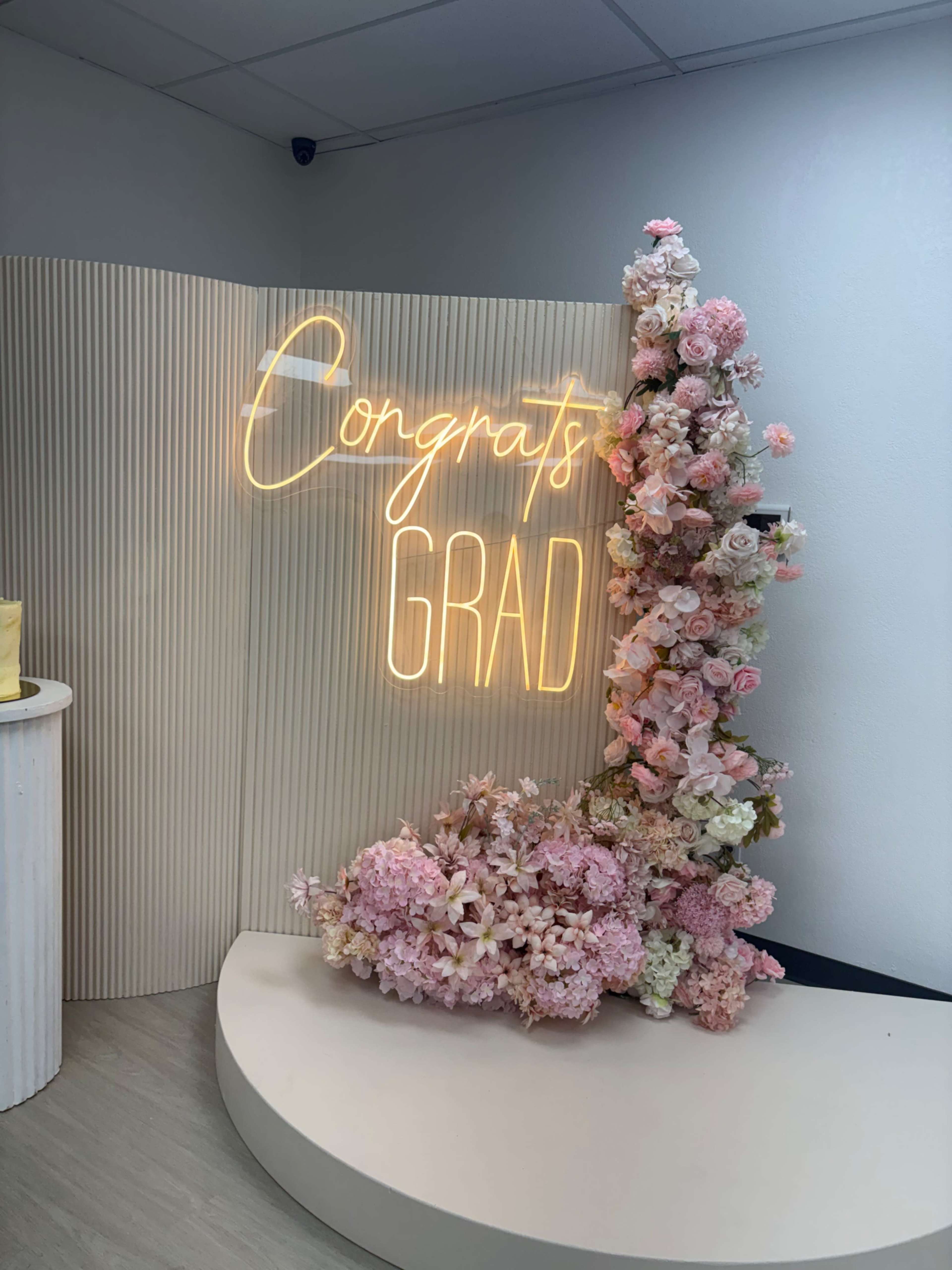The image shows a decorative setup featuring a "Congrats Grad" neon sign surrounded by pink and white flowers on a curved backdrop.