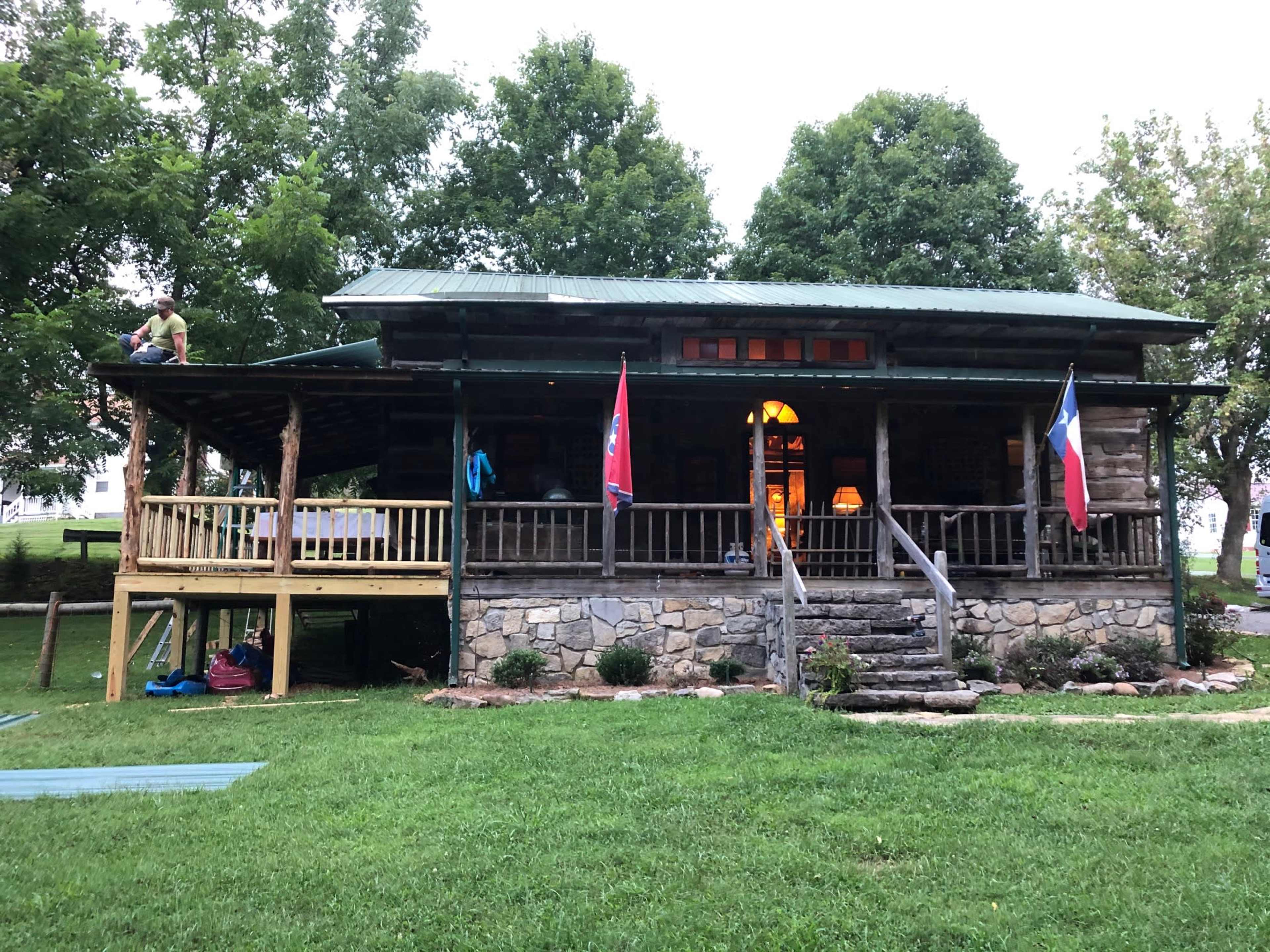 A wooden cabin with a green roof features a porch, stone steps, and flags from Texas and Tennessee, surrounded by grassy areas and trees.