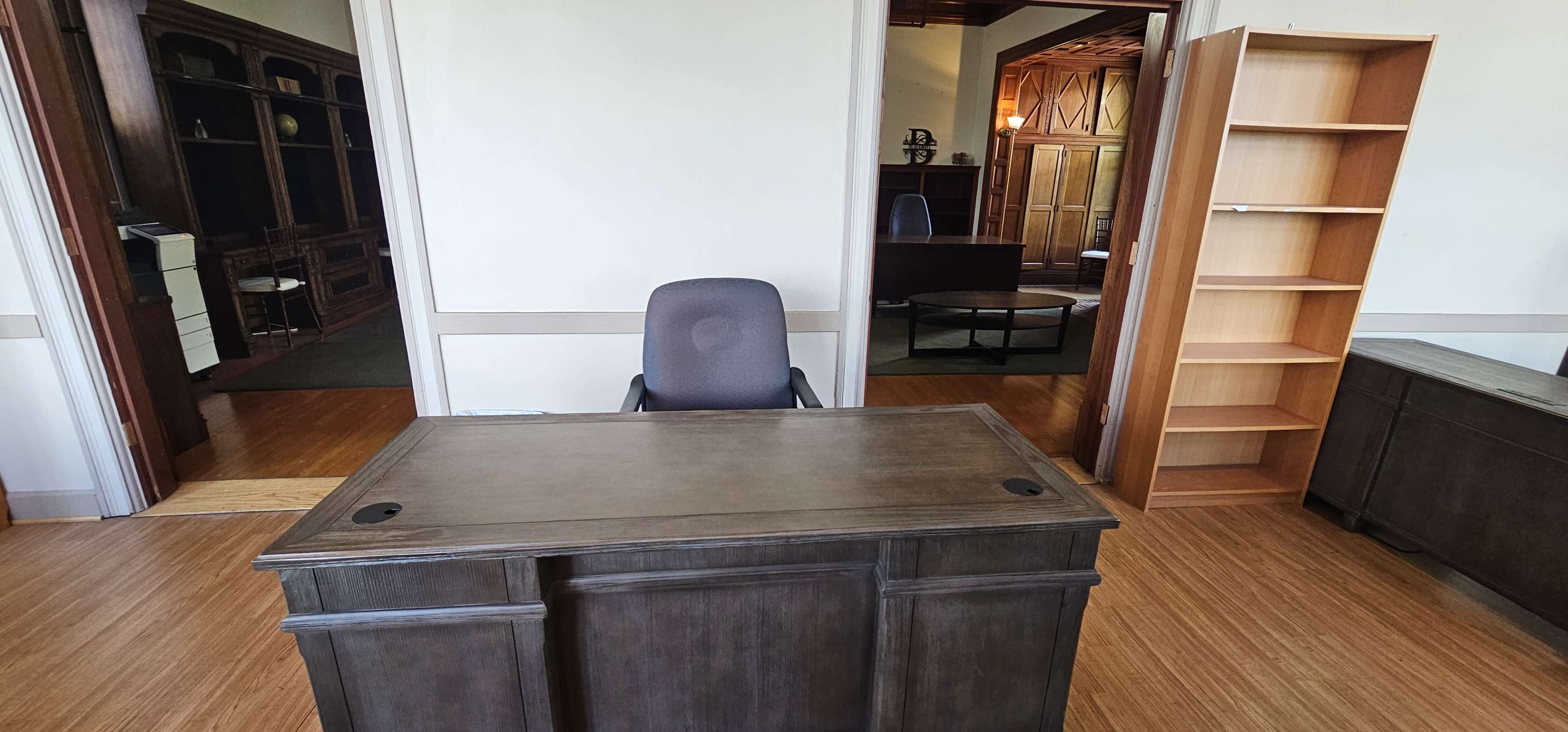 The image shows a sparse office space featuring a wooden desk with an office chair in front of it, and a bookshelf on the right.