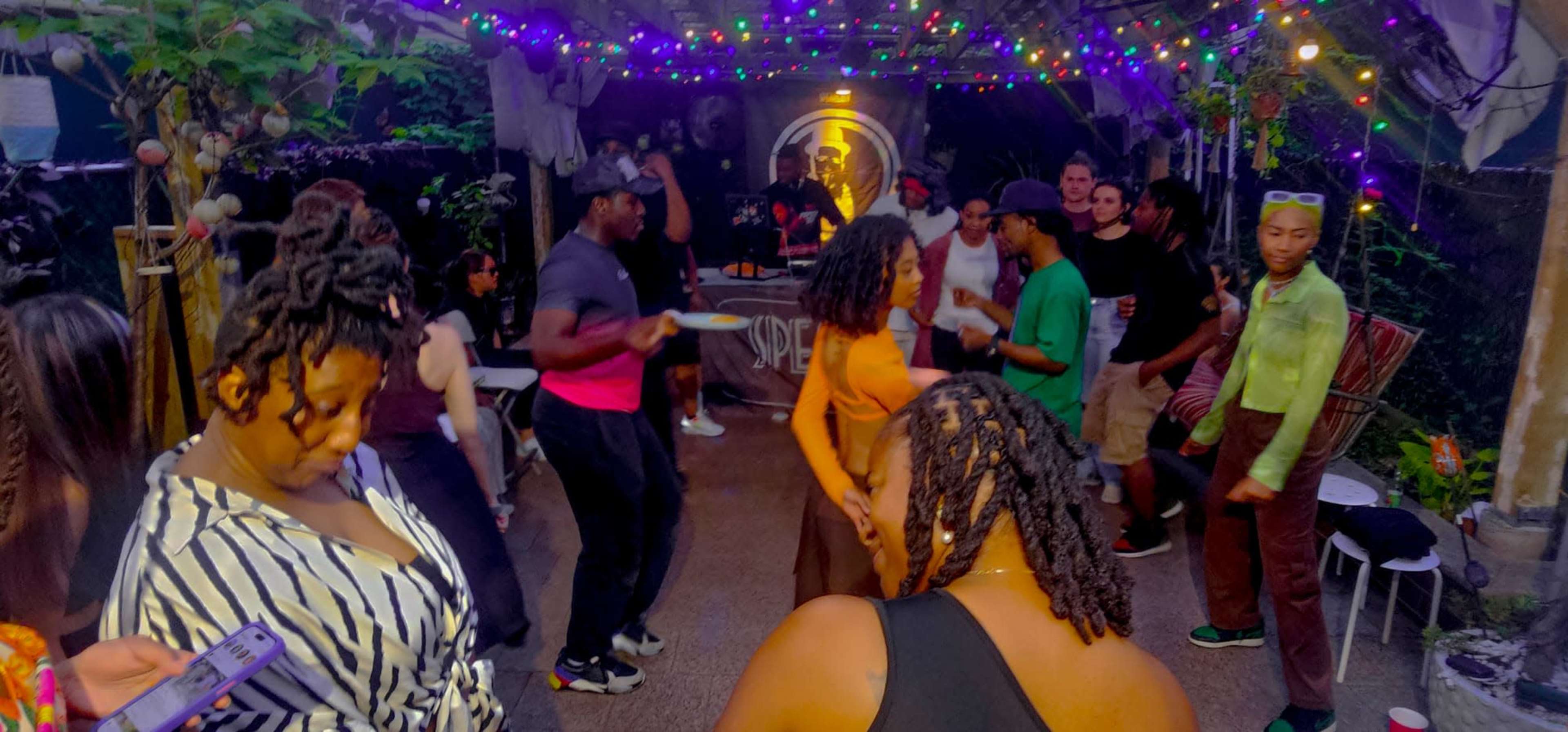 A group of people is dancing and socializing at an outdoor party decorated with colorful lights.