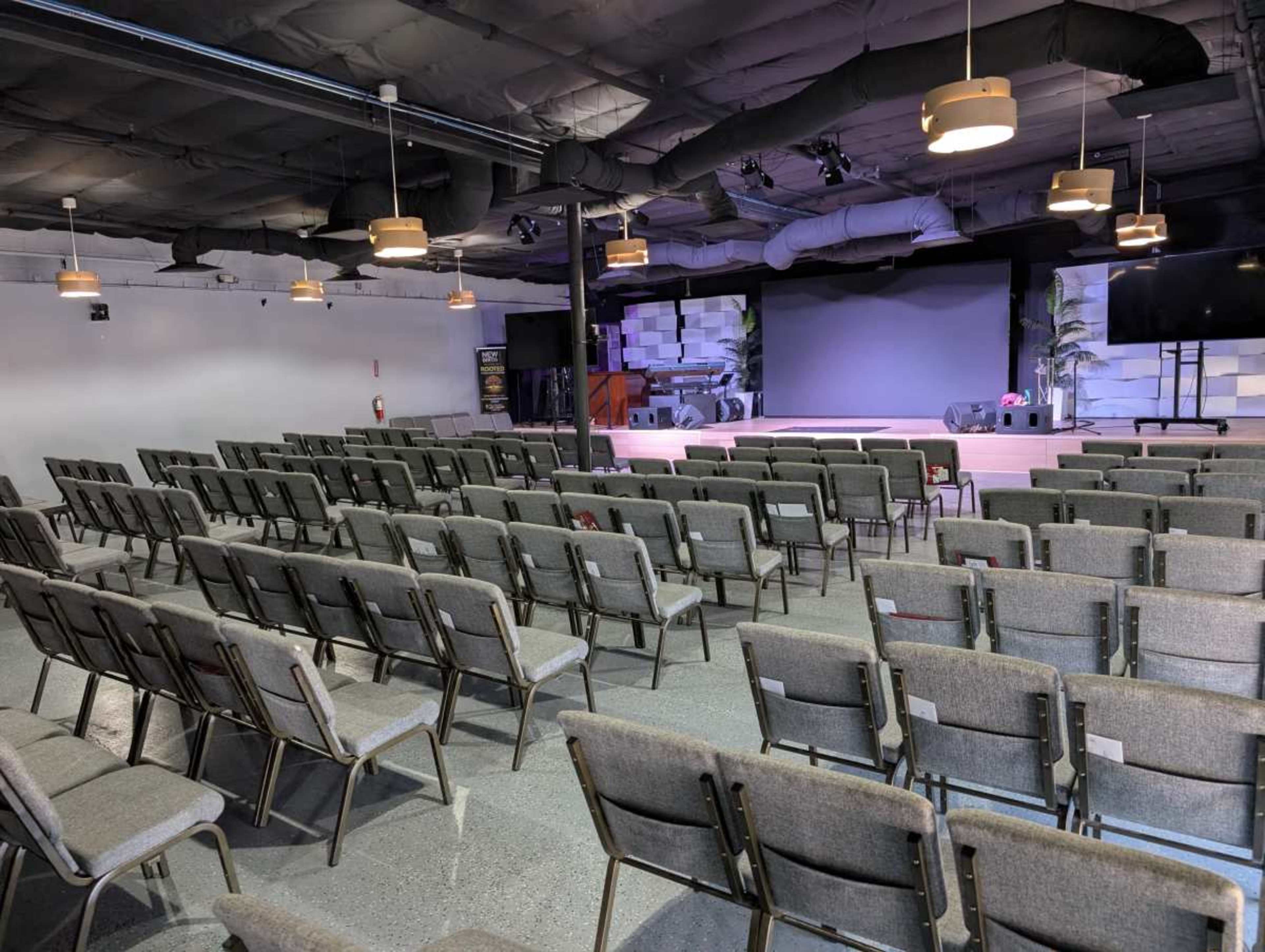 The image shows an empty auditorium with rows of gray chairs facing a stage that has a large screen and is flanked by two plants.