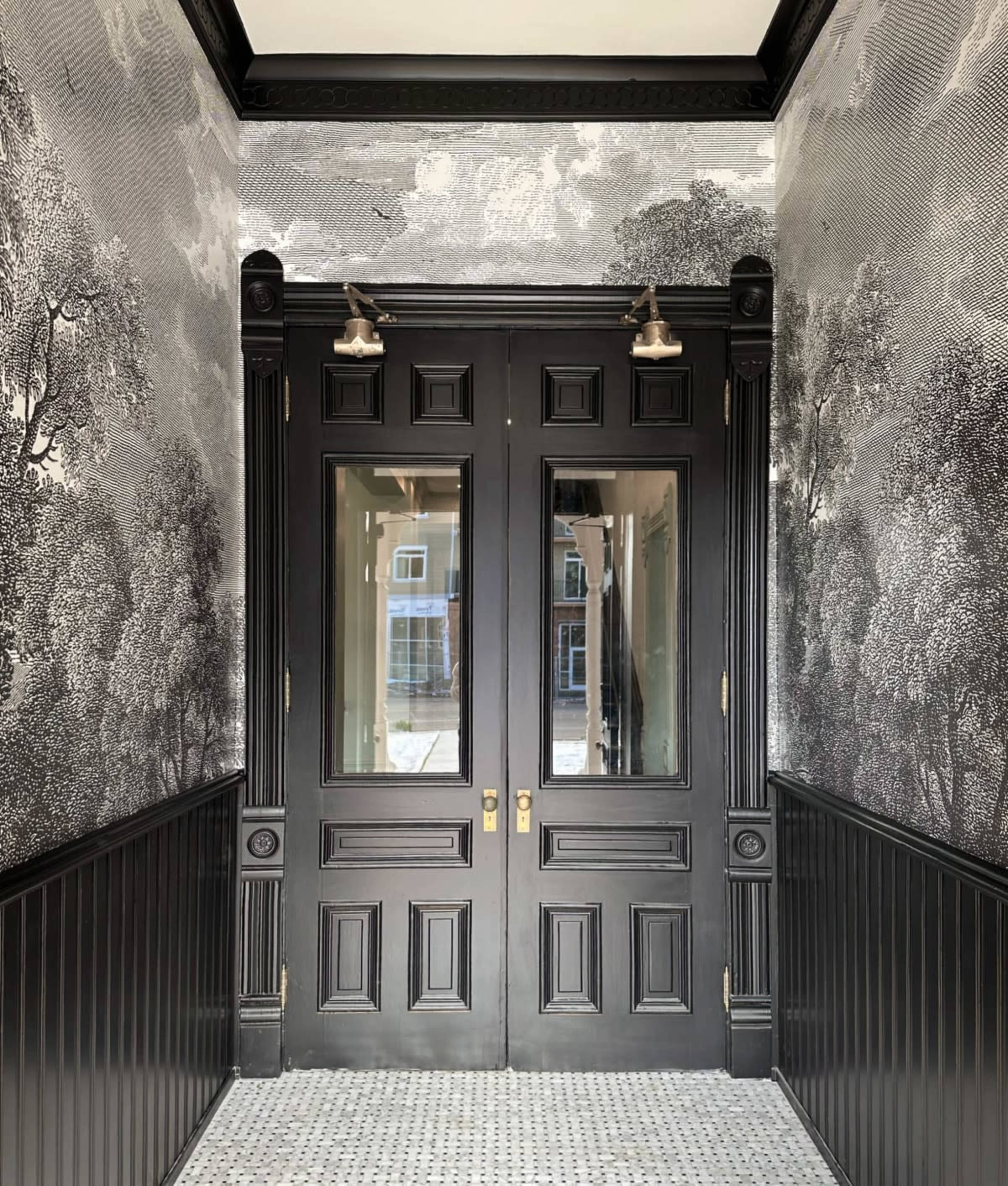 A narrow hallway featuring black double doors surrounded by detailed wallpaper of trees, with wall sconces illuminating the space.