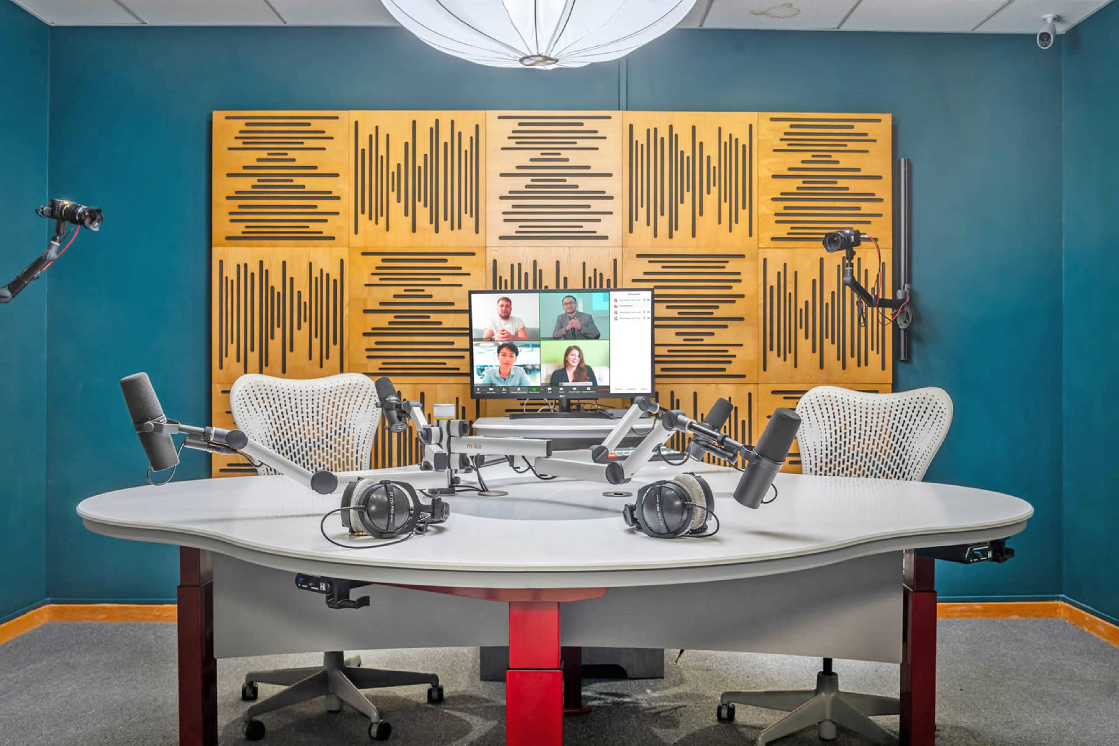 The image shows a modern recording studio with two chairs, multiple microphones, headphones, and a display screen showing remote participants.