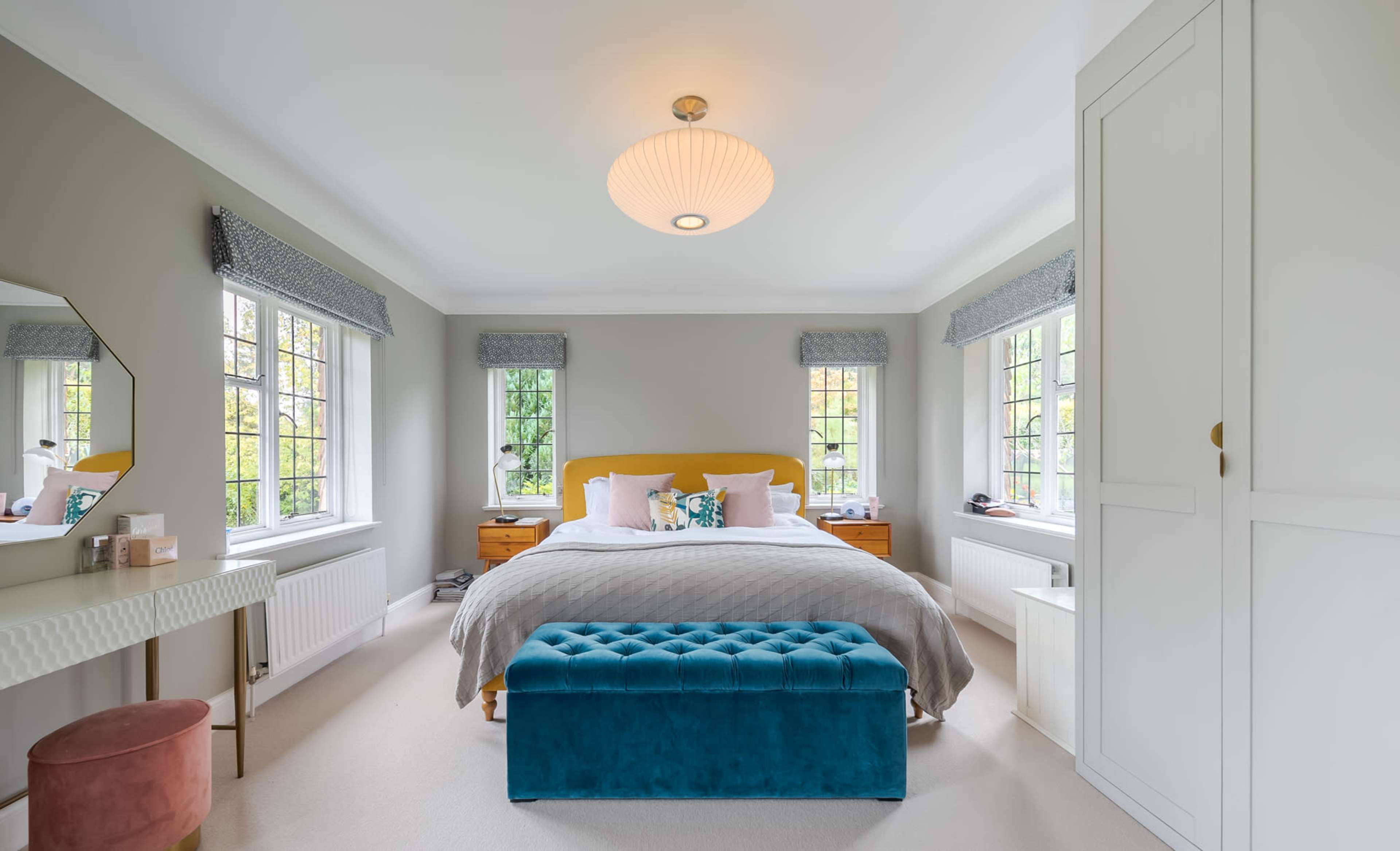 A spacious bedroom features a large bed with a gray quilt, a teal storage bench, and dressed windows beside wooden nightstands.