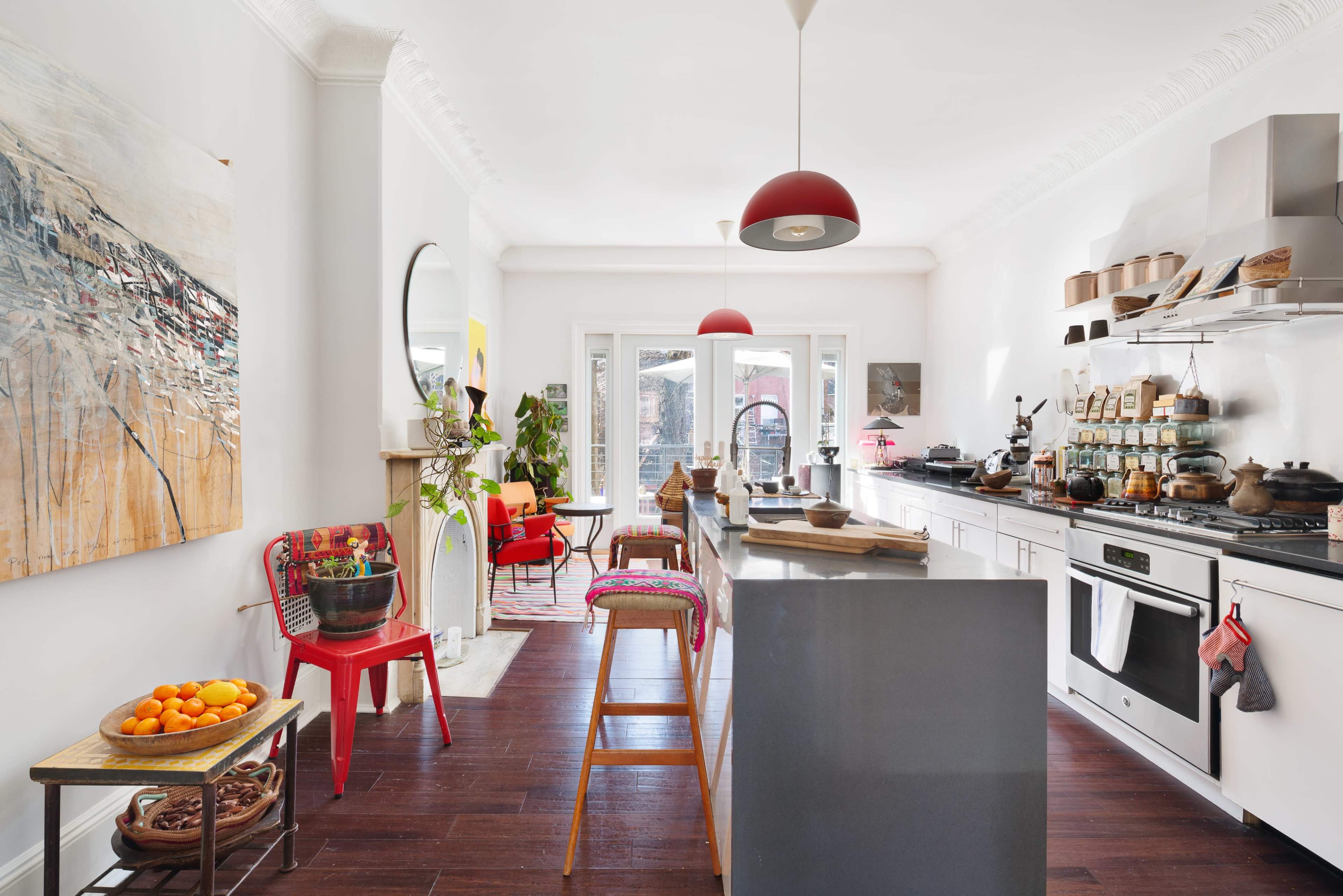 A bright kitchen features a central island, red pendant lights, and a mix of modern and vintage decor, with pots and ingredients displayed on shelves and a sitting area visible through the windows.