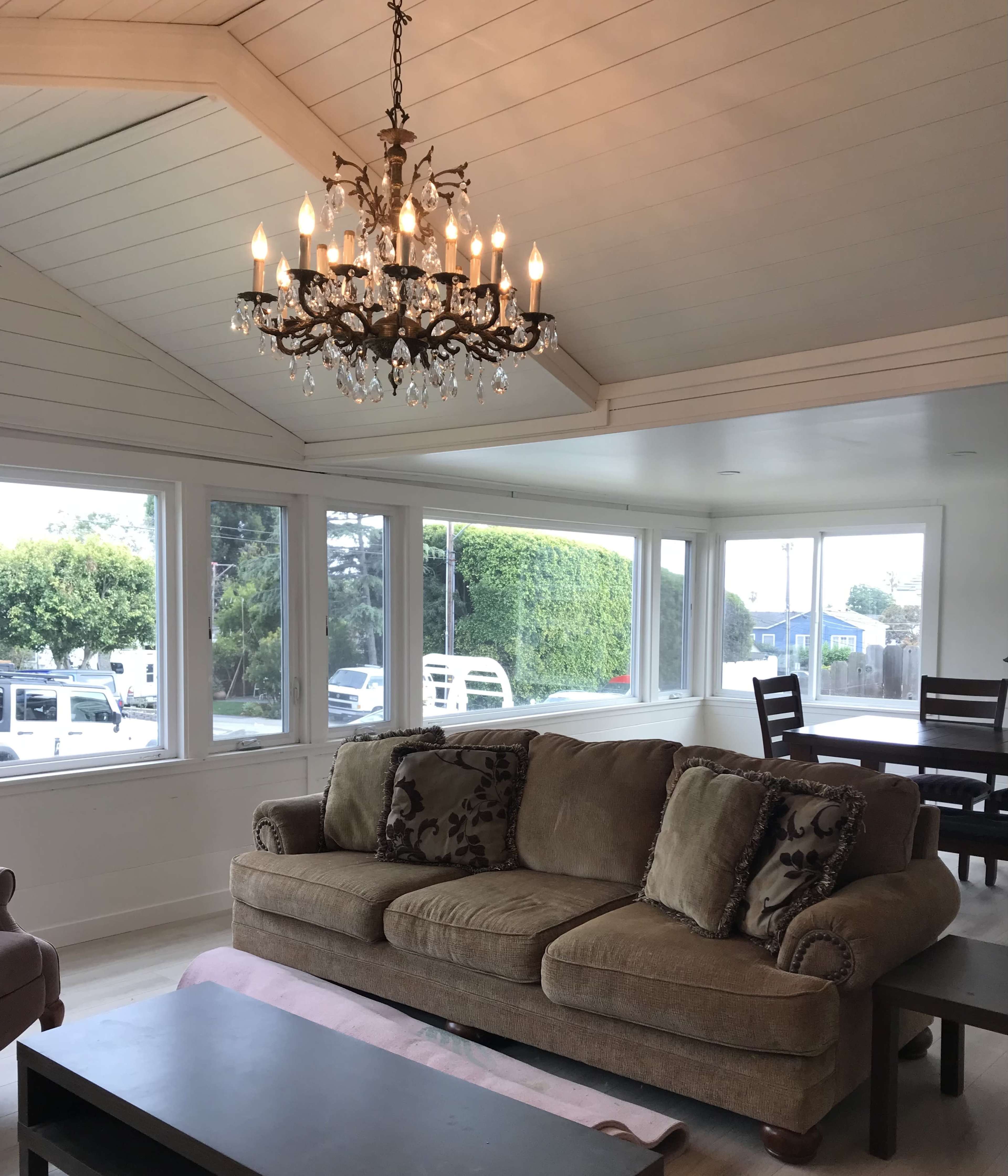 A living room with a large chandelier hanging from a white, vaulted ceiling, a beige sofa, and windows that offer a view of the outdoors.