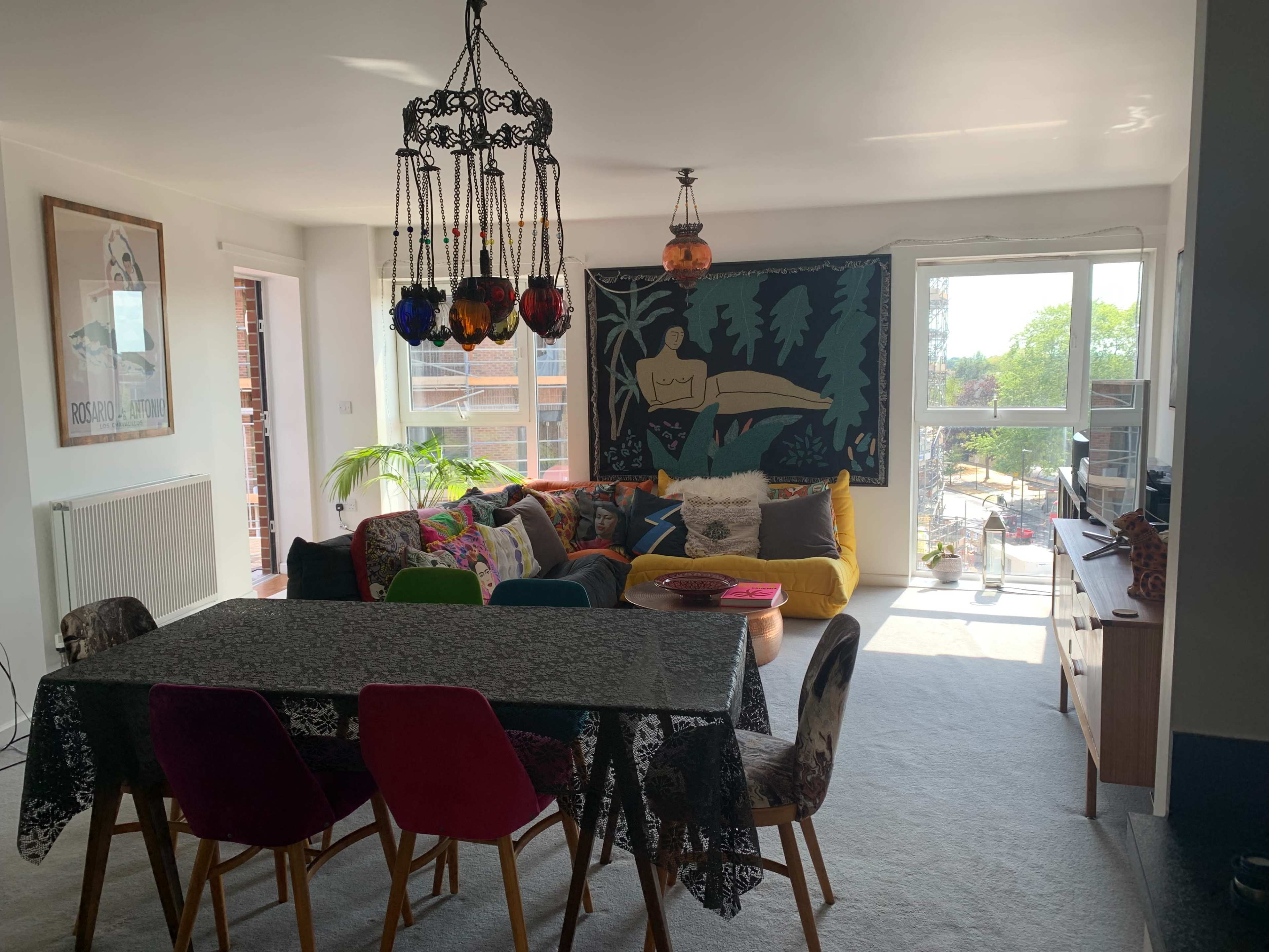 The room features a bright living area with a colorful sofa, a dining table with upholstered chairs, and large windows providing natural light.