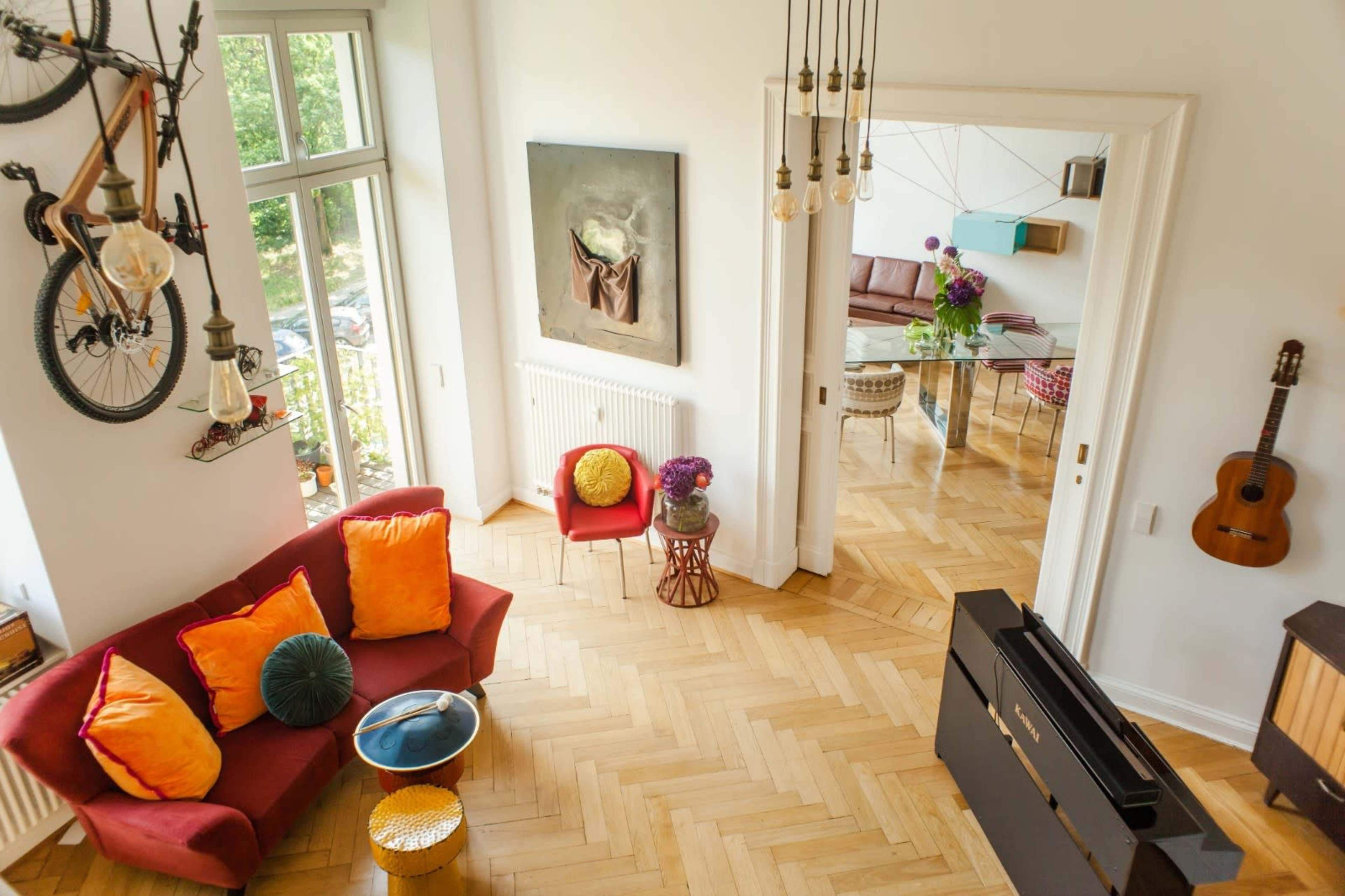 A bright living area features a red sofa with colorful cushions, a wooden floor, and a guitar on the wall, complemented by large windows and a dining space in the background.