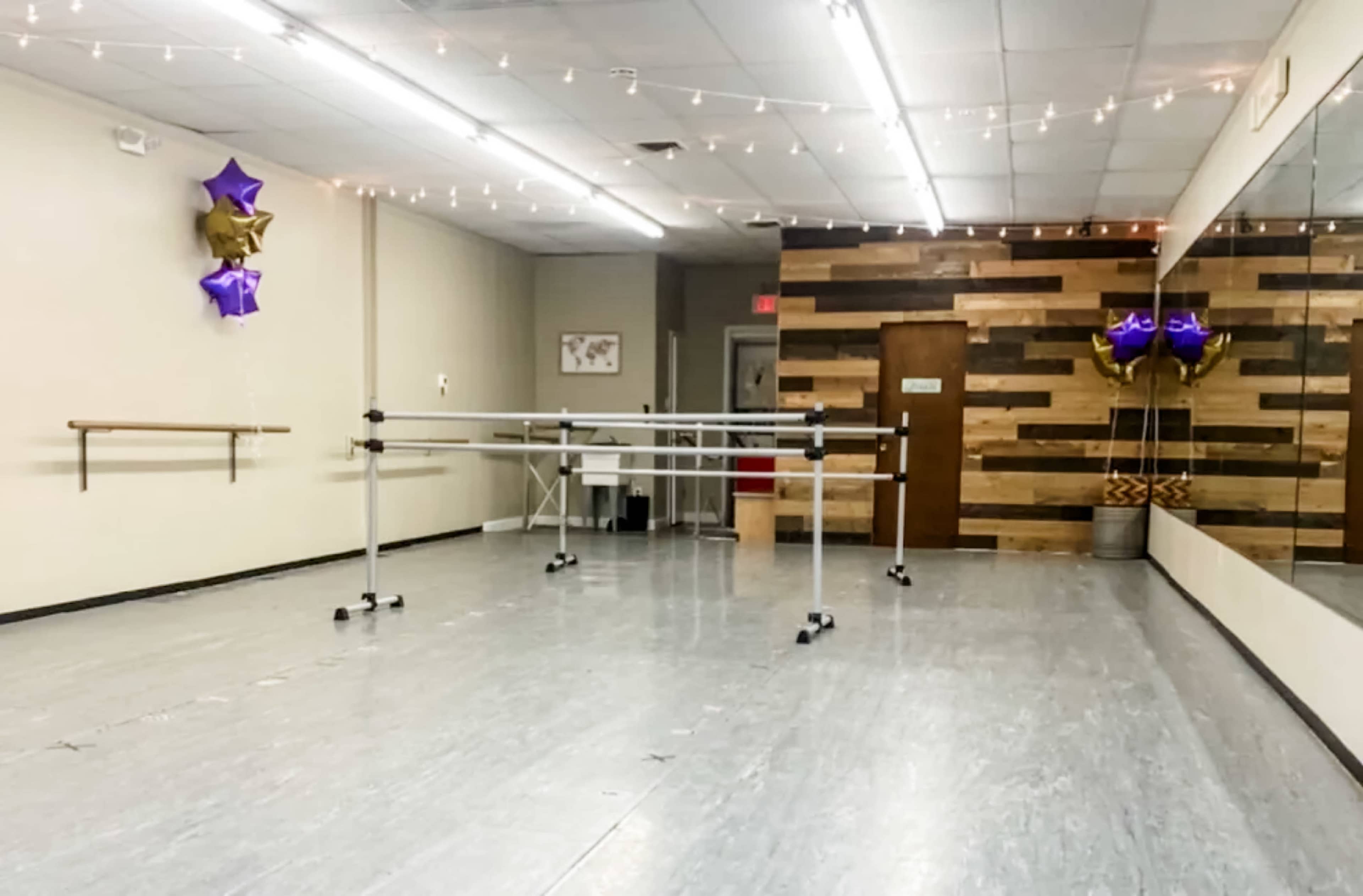 The image shows a spacious dance studio with mirrored walls, wooden accents, and ballet bars along one side, decorated with balloons and string lights.