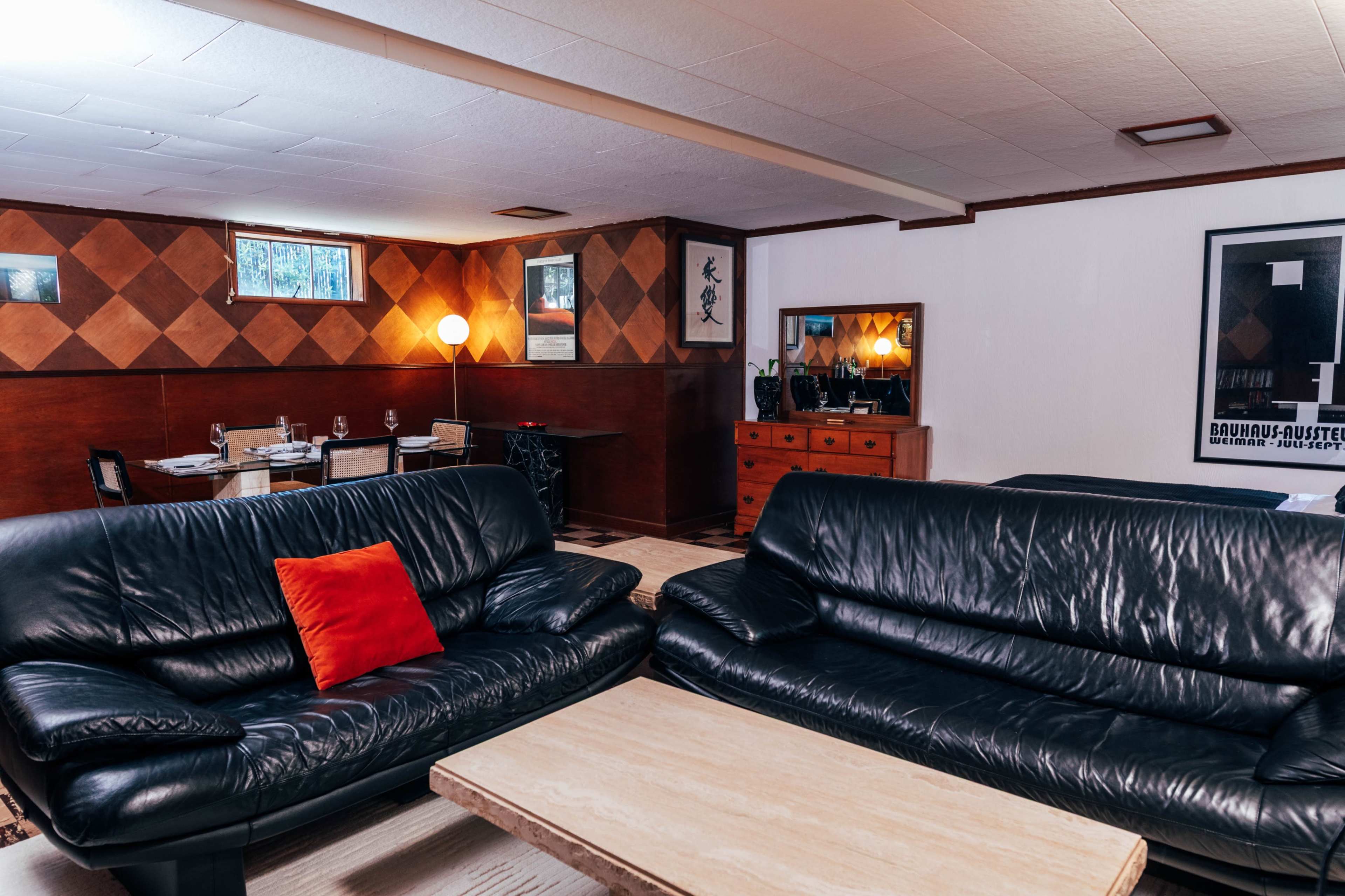 The image shows a spacious living area featuring two black leather sofas and a wooden coffee table, with a dining area visible in the background.