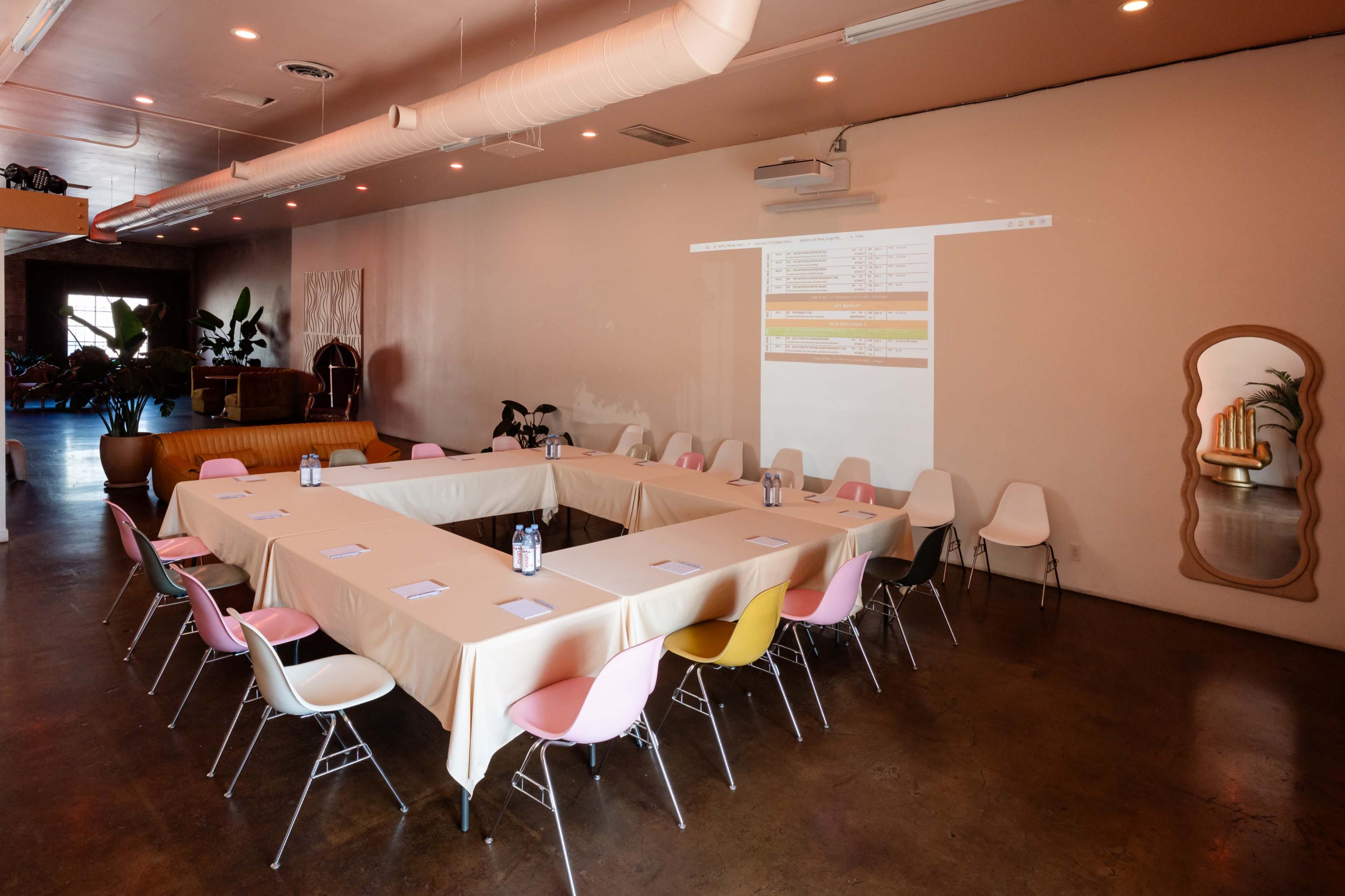 A meeting room is arranged with a rectangular table covered by a beige tablecloth, surrounded by colorful chairs, and a screen displaying information on the wall.