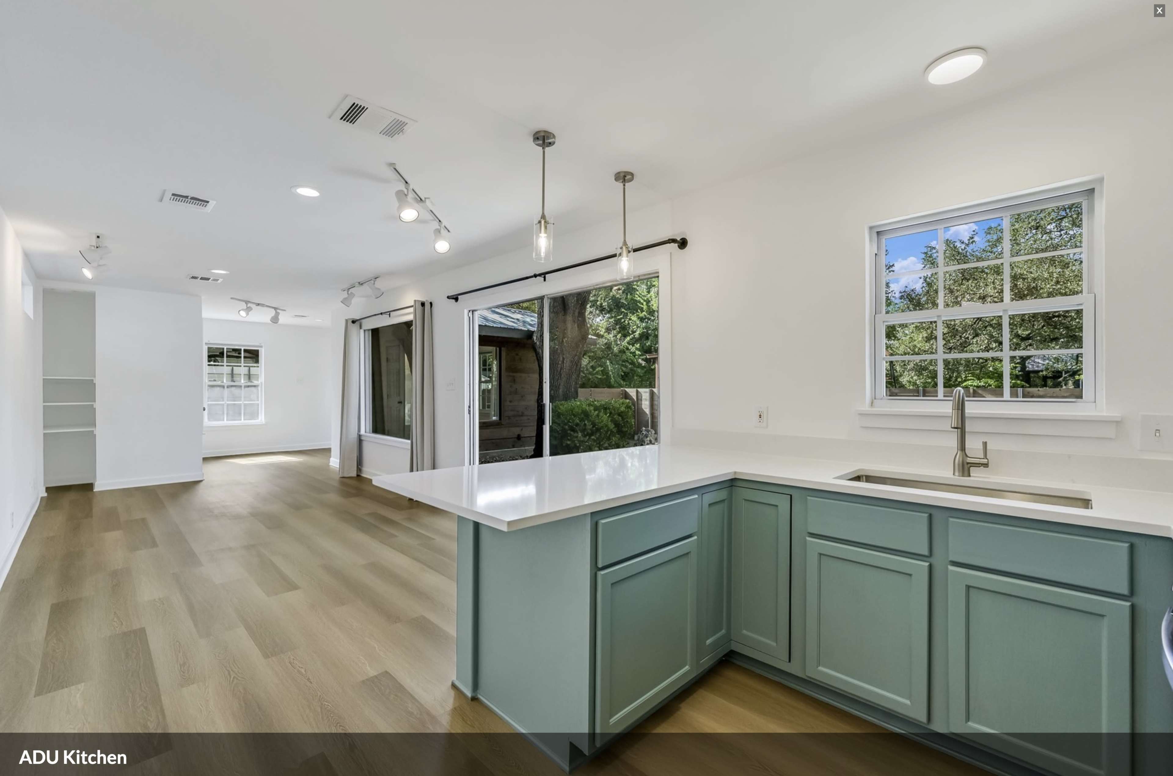 The image shows a modern kitchen with light-colored cabinets, a large window, and an open layout leading to a patio area.