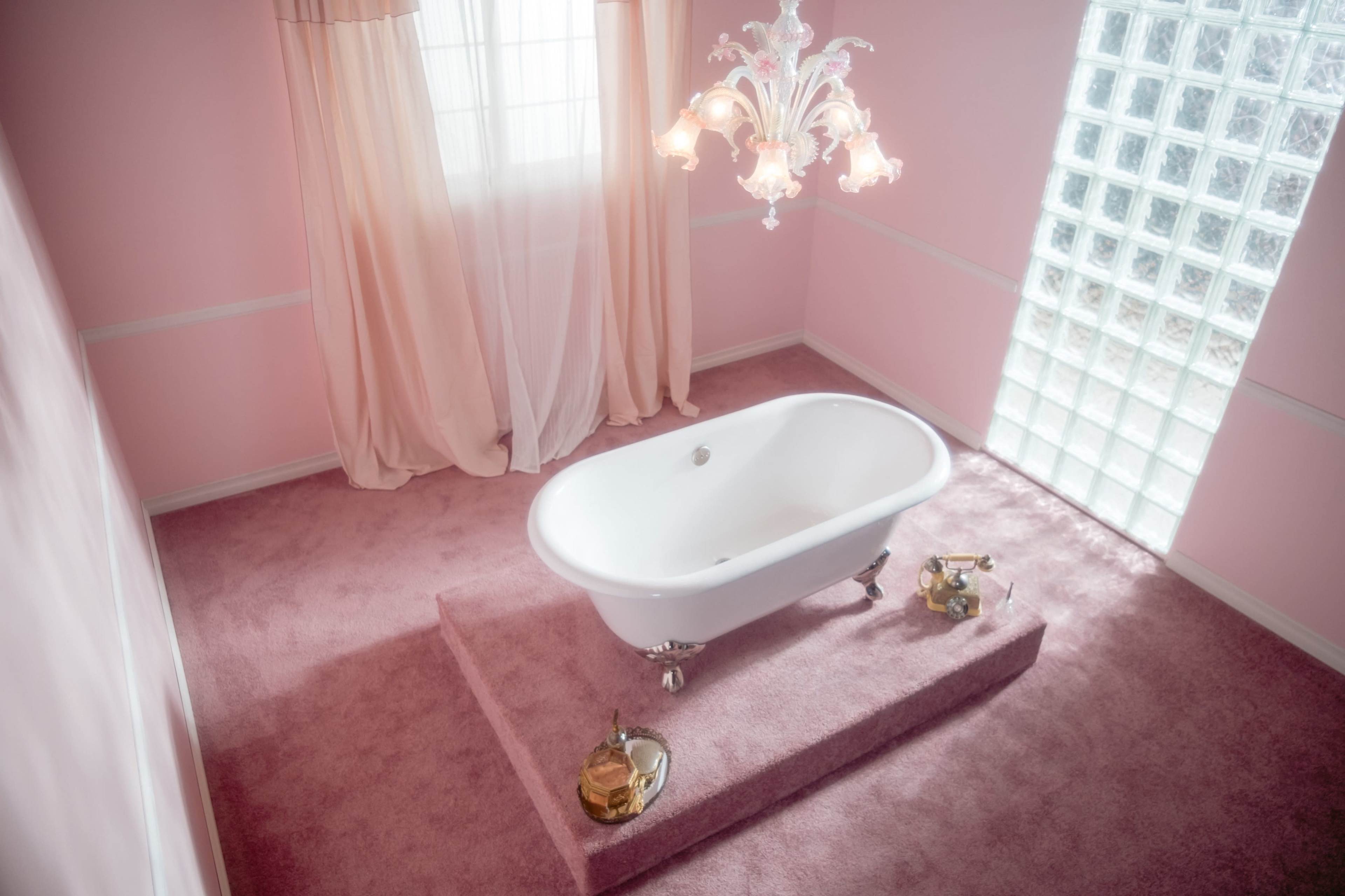 A white freestanding bathtub is situated on a raised platform in a pink, softly lit bathroom with a chandelier and glass block window.