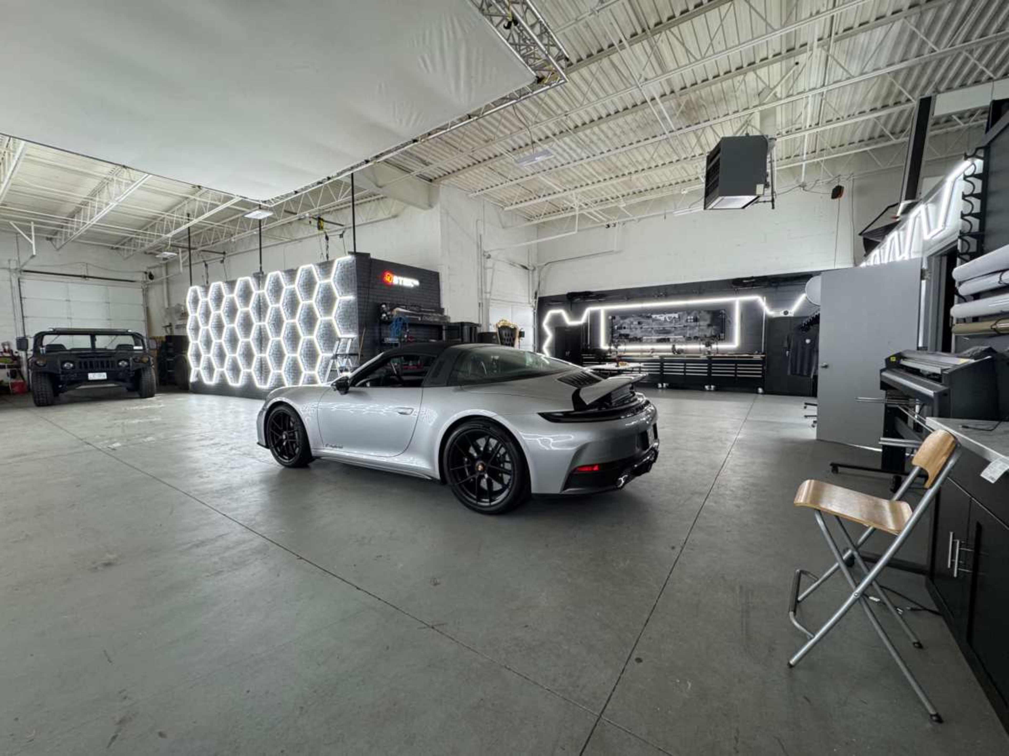 A silver sports car is parked in a spacious garage featuring modern lighting and various vehicles along the walls.