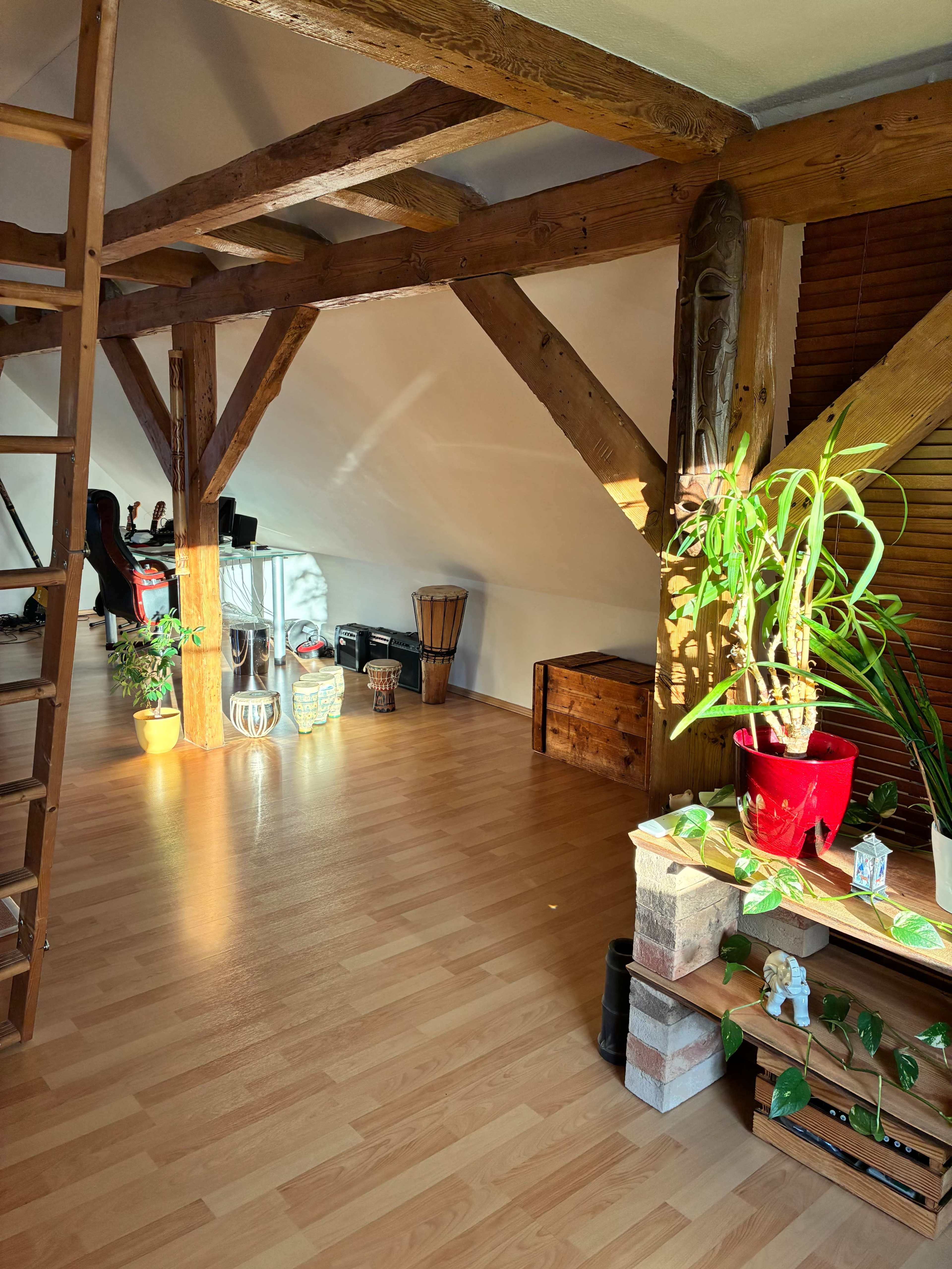 The image shows a spacious loft with wooden beams, a staircase, and plants, featuring various furniture items including a drum, a potted plant, and a wooden storage box.