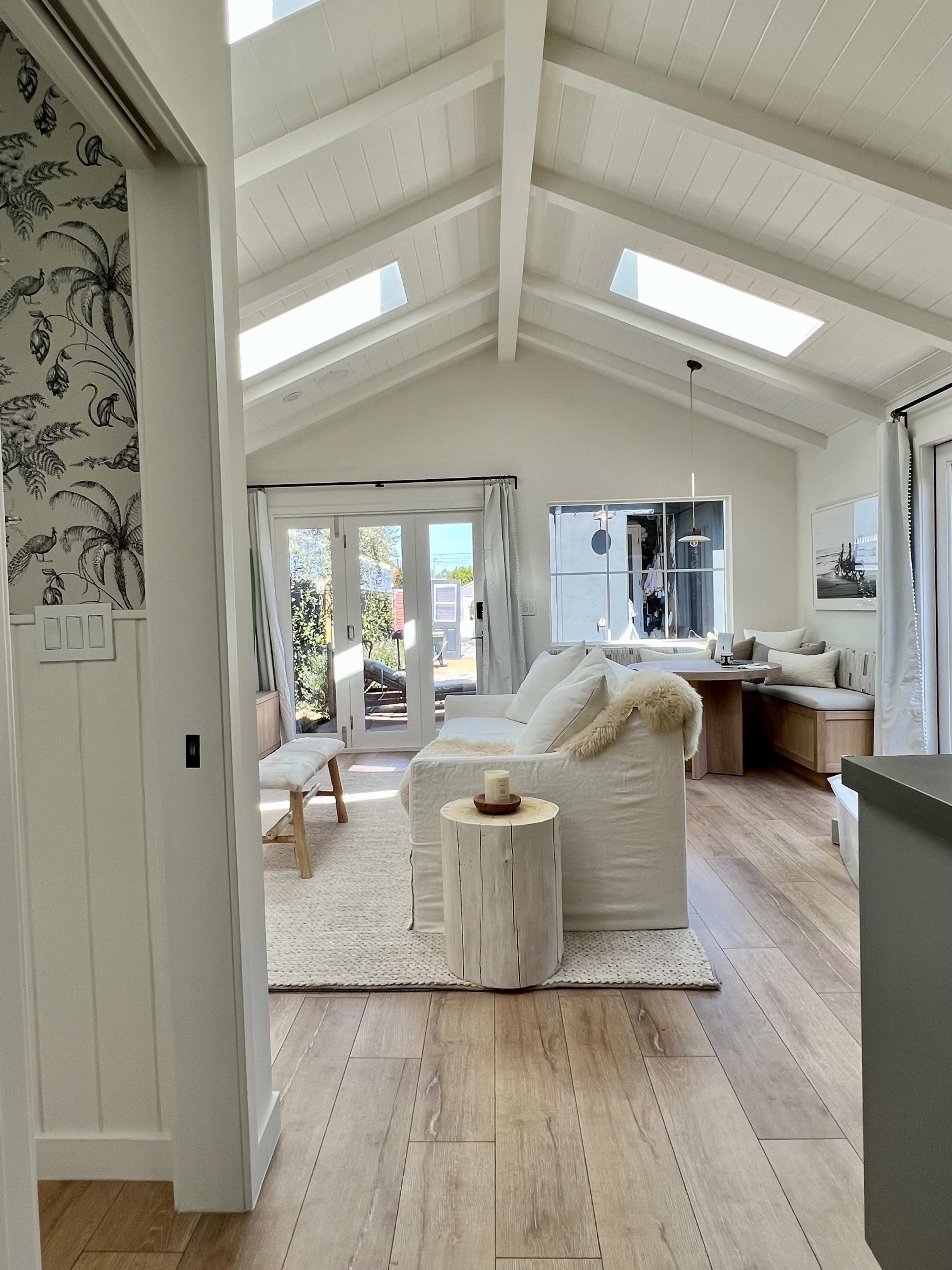 A bright, open living area with a high ceiling, featuring a white sofa, wooden furniture, and large windows that let in natural light.
