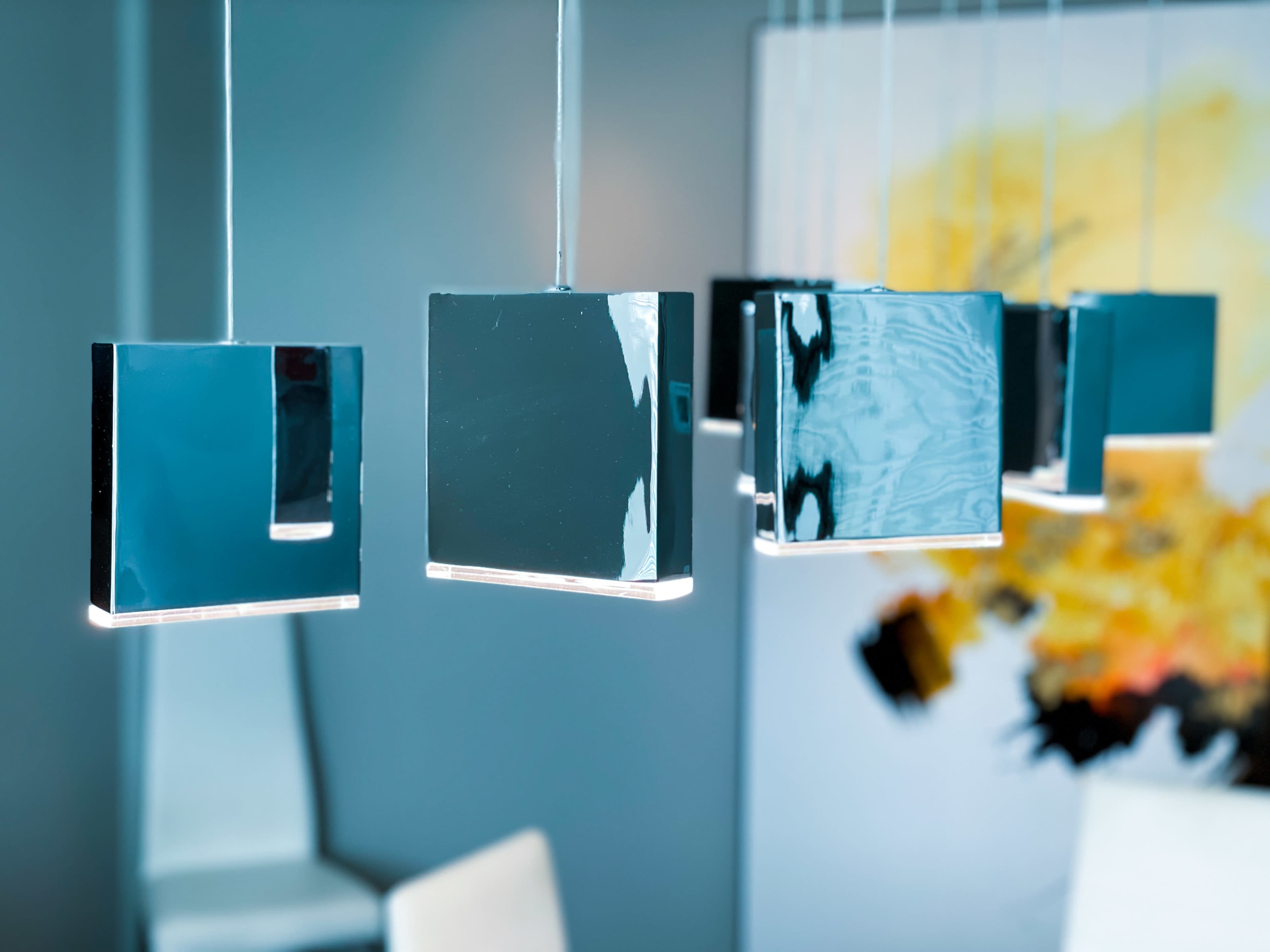 Several modern, square hanging light fixtures above a table, with an abstract yellow and black painting in the background.