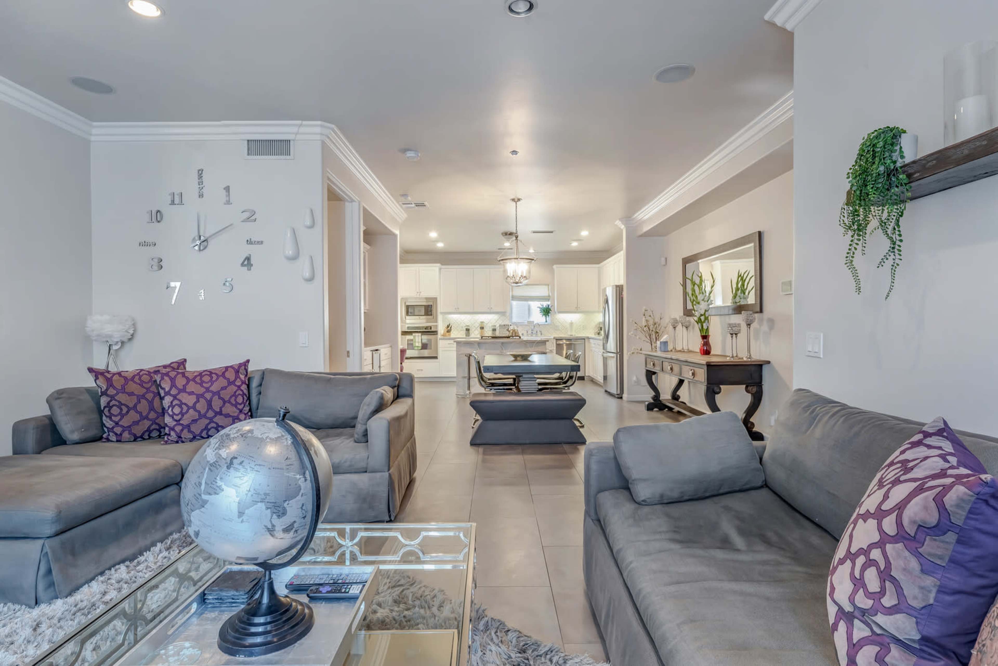 A modern, open-concept living room and kitchen area featuring grey sofas, decorative pillows, a glass coffee table, and a wall clock.