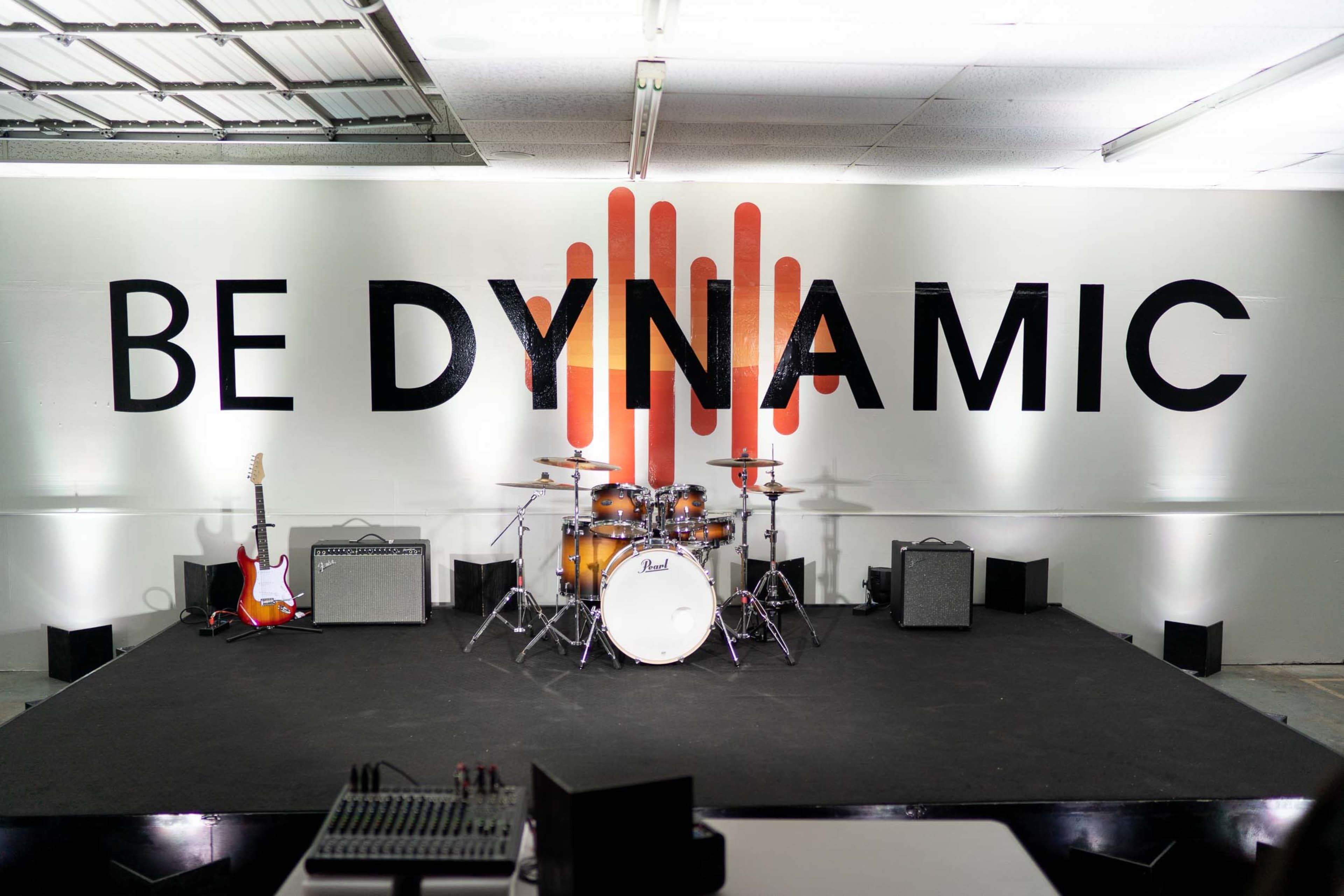 A stage setup with a drum kit, guitars, amplifiers, and a large wall displaying the text "BE DYNAMIC" in bold letters.