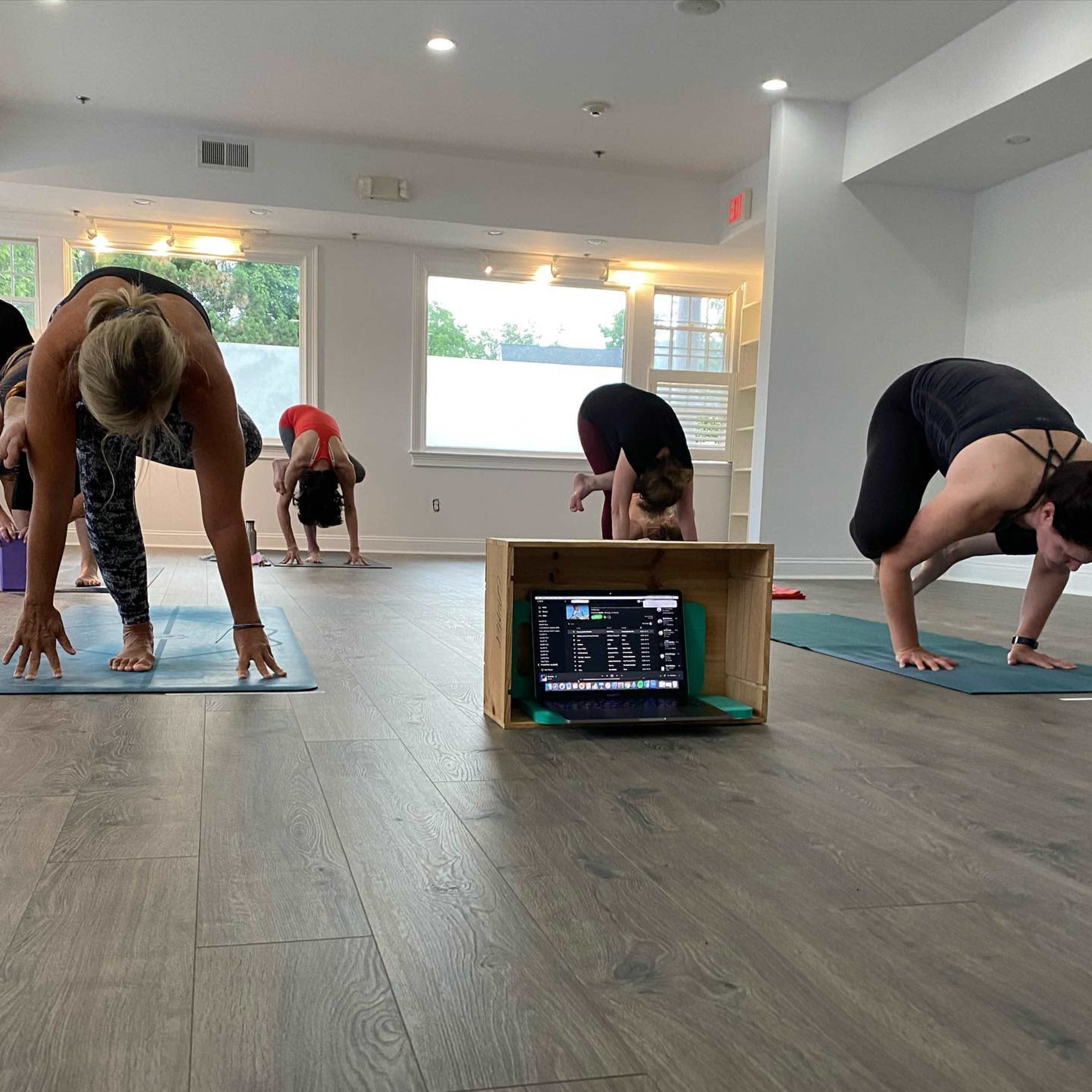 A group of individuals practices yoga in a bright, spacious studio with a laptop placed on a wooden crate in the foreground.
