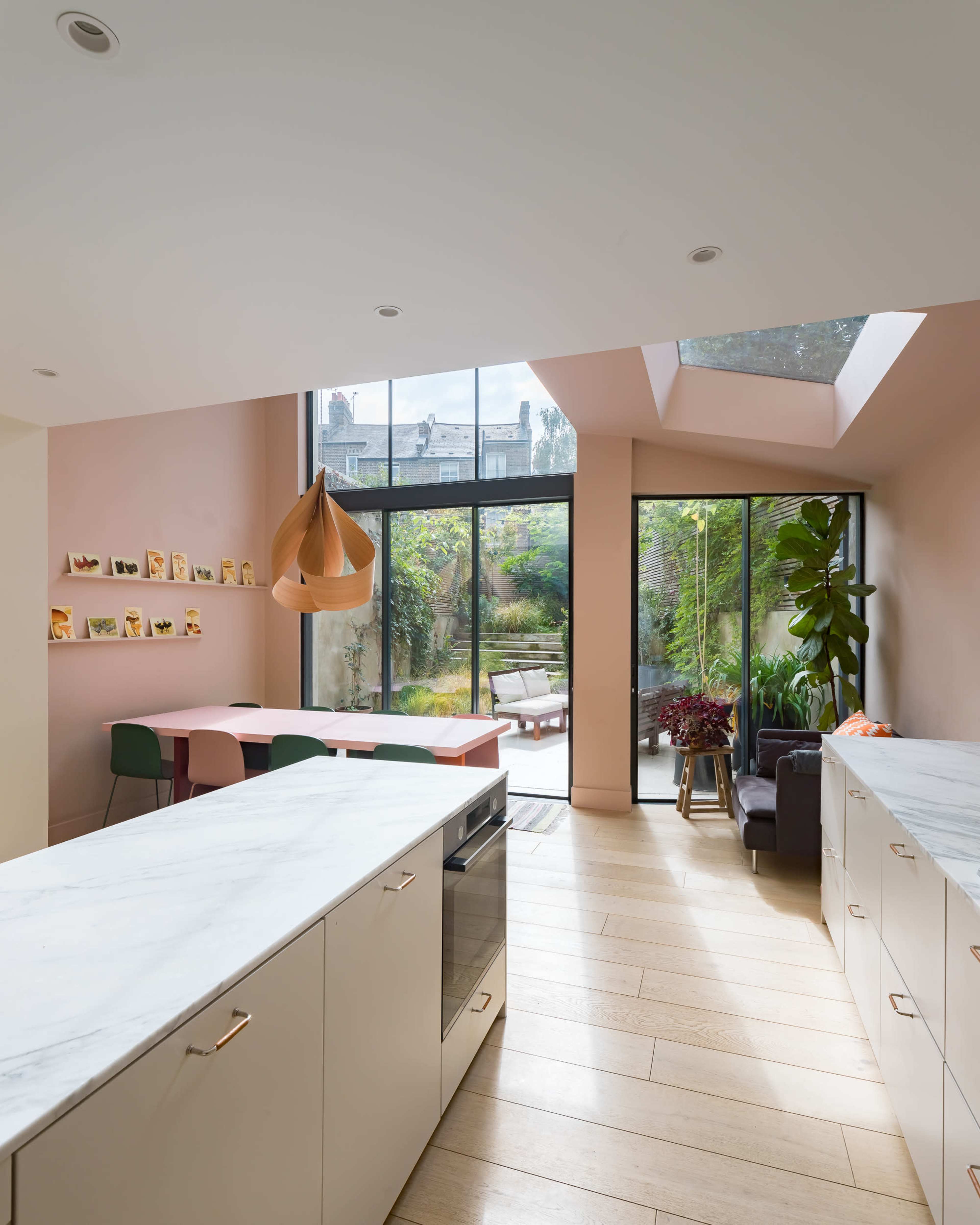 A modern kitchen features marble countertops, light-colored cabinetry, and large windows overlooking a garden.