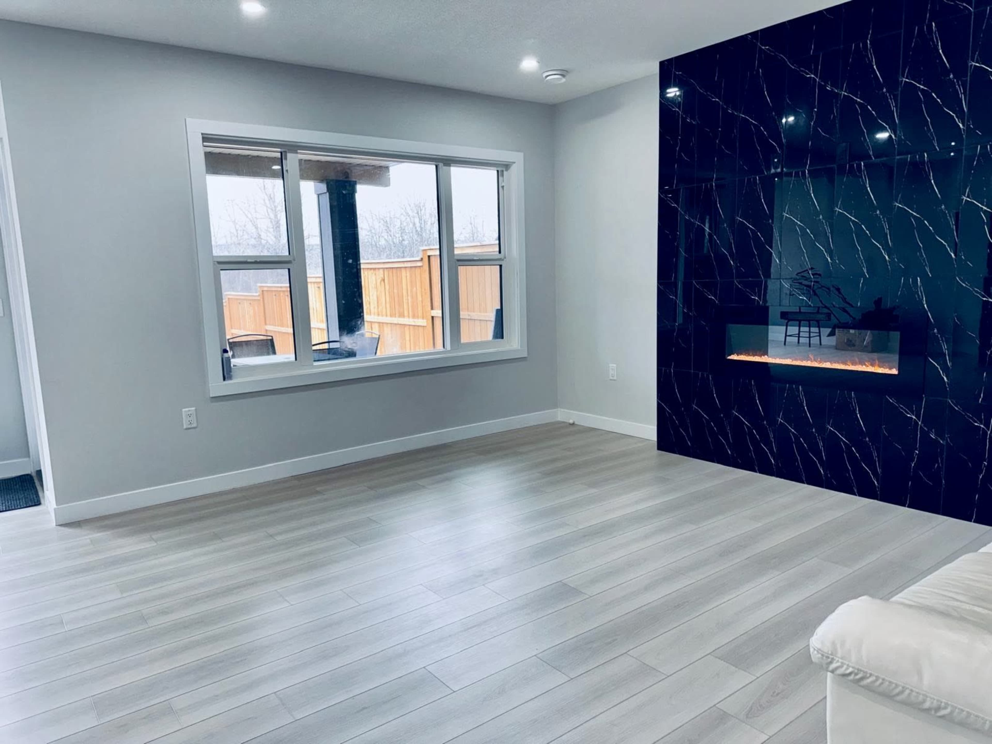 The image shows a modern living room with light-colored wooden flooring, a large window, and a stylish black feature wall with an electric fireplace.