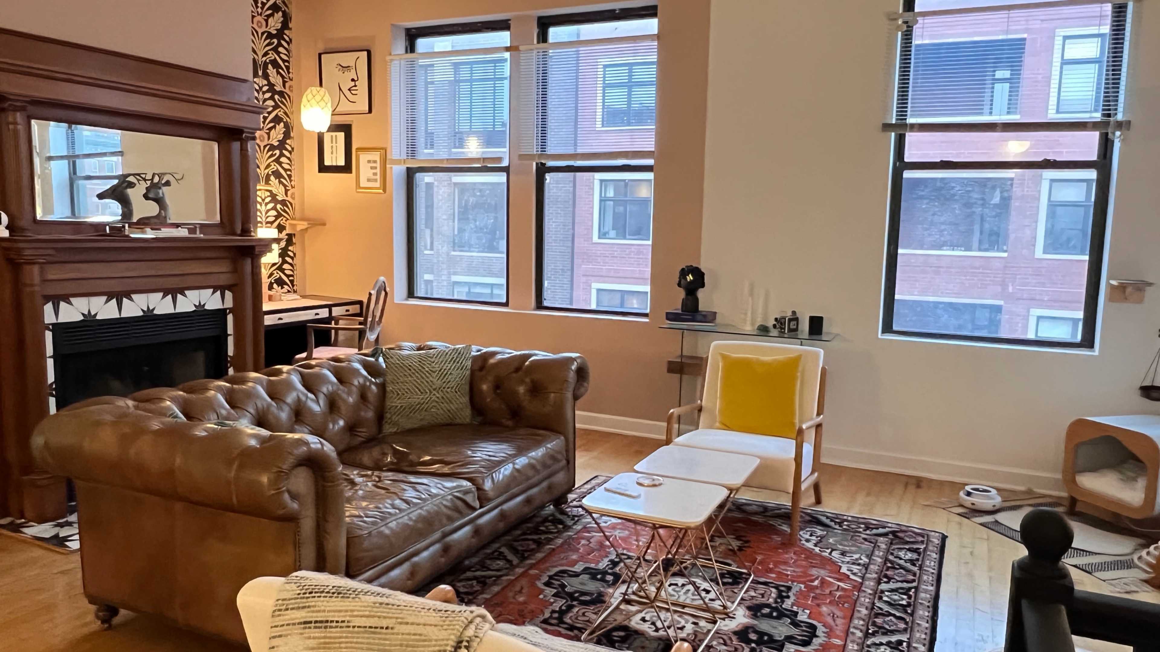 A cozy living room with a brown leather chesterfield sofa, a white armchair, a coffee table, and a patterned area rug, along with large windows letting in natural light.