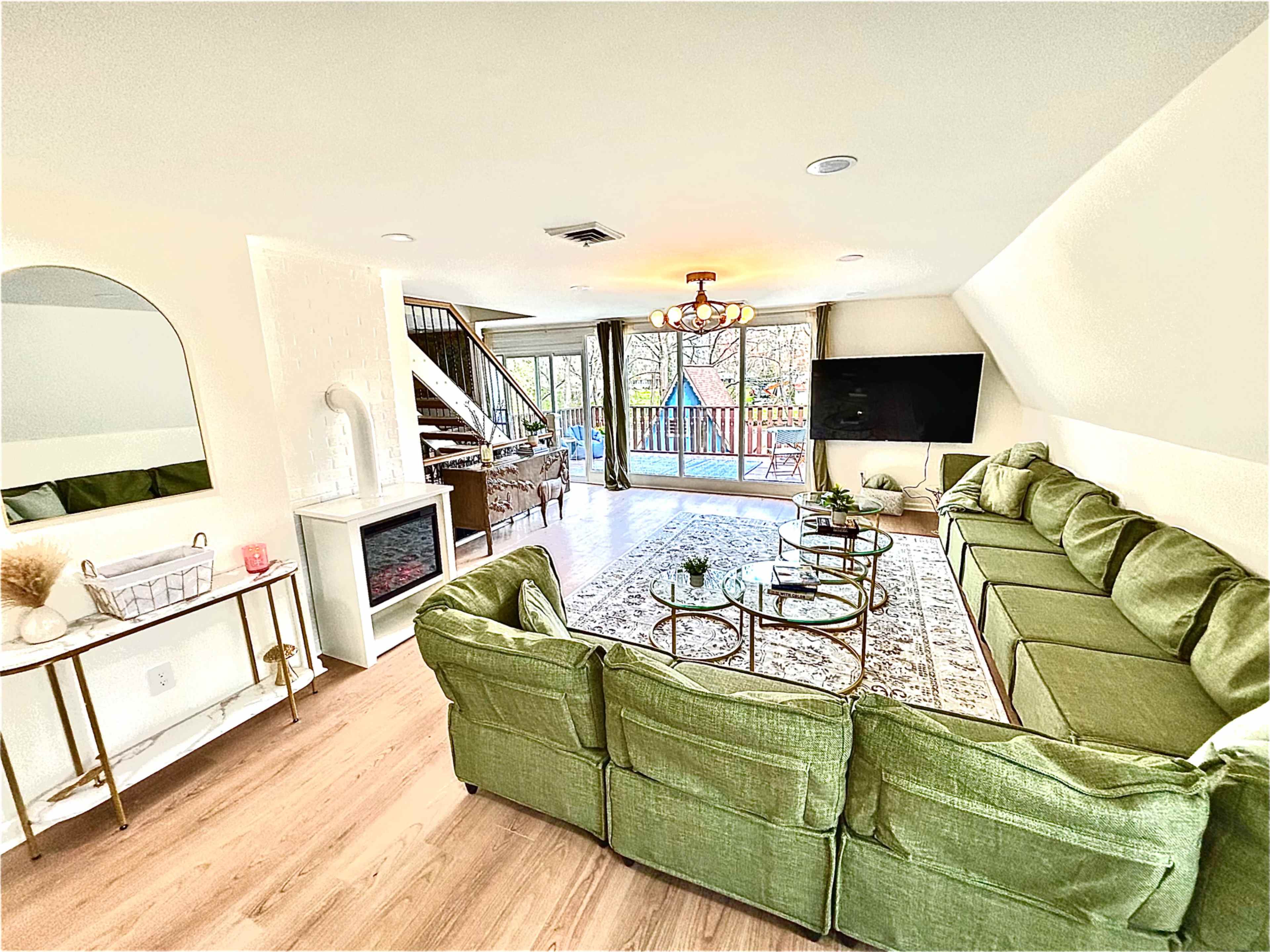 A spacious living room furnished with a large green sectional sofa, a glass coffee table, and a flat-screen TV, with large windows providing a view of the outdoors.
