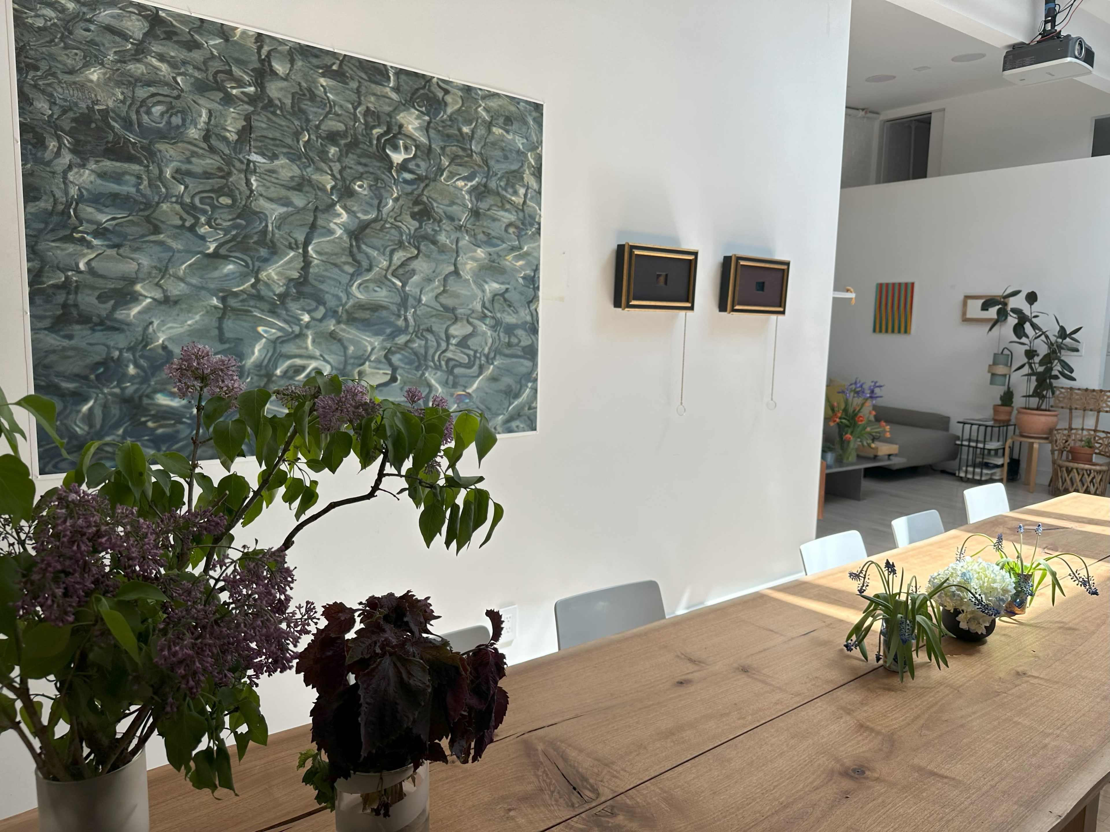 A bright, modern dining area with a wooden table featuring potted plants, a wall art piece depicting water, and a small decorative arrangement on the right.
