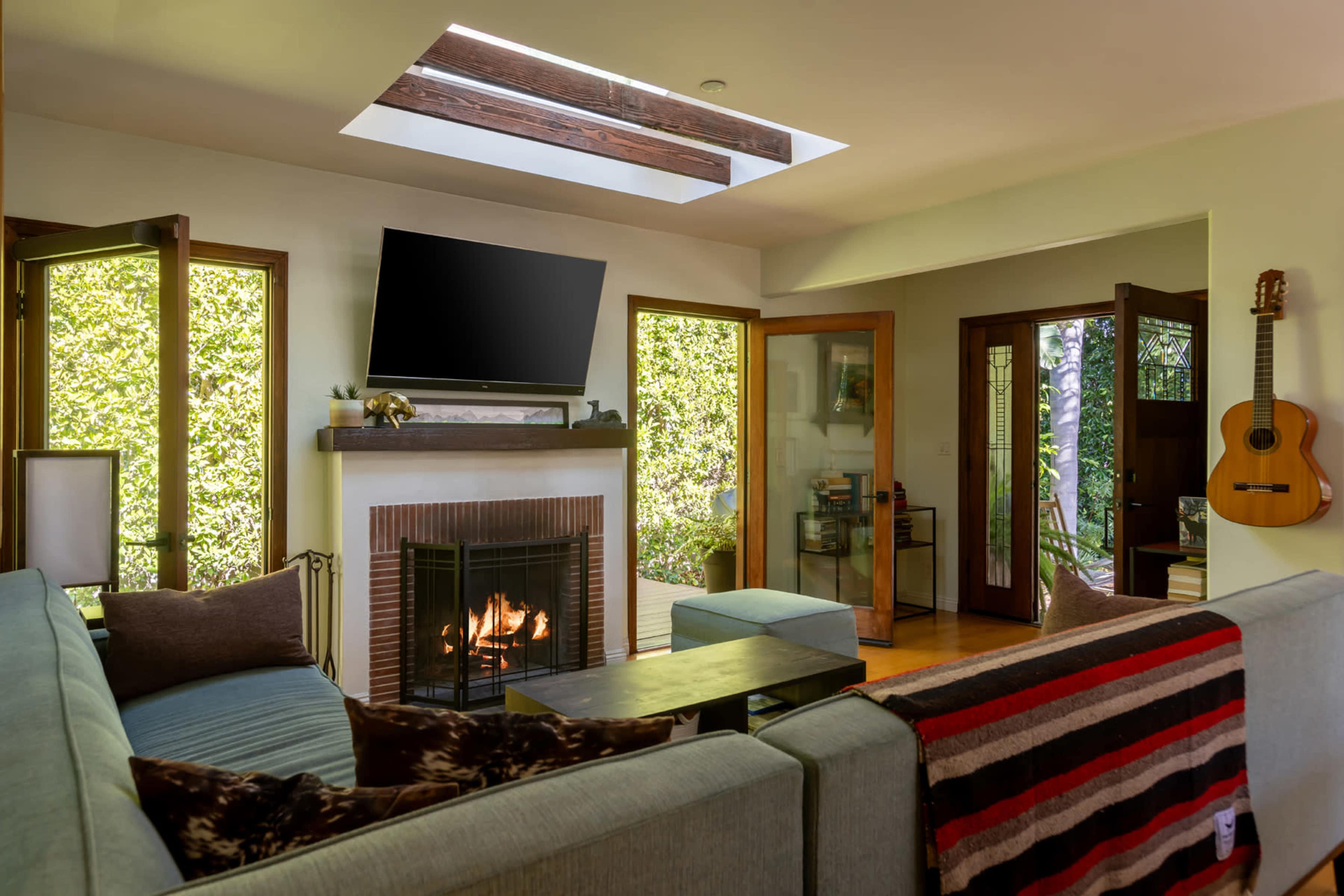 The image shows a cozy living room with a fireplace, large windows, and a skylight, featuring a television mounted above the mantel and a guitar hanging on the wall.