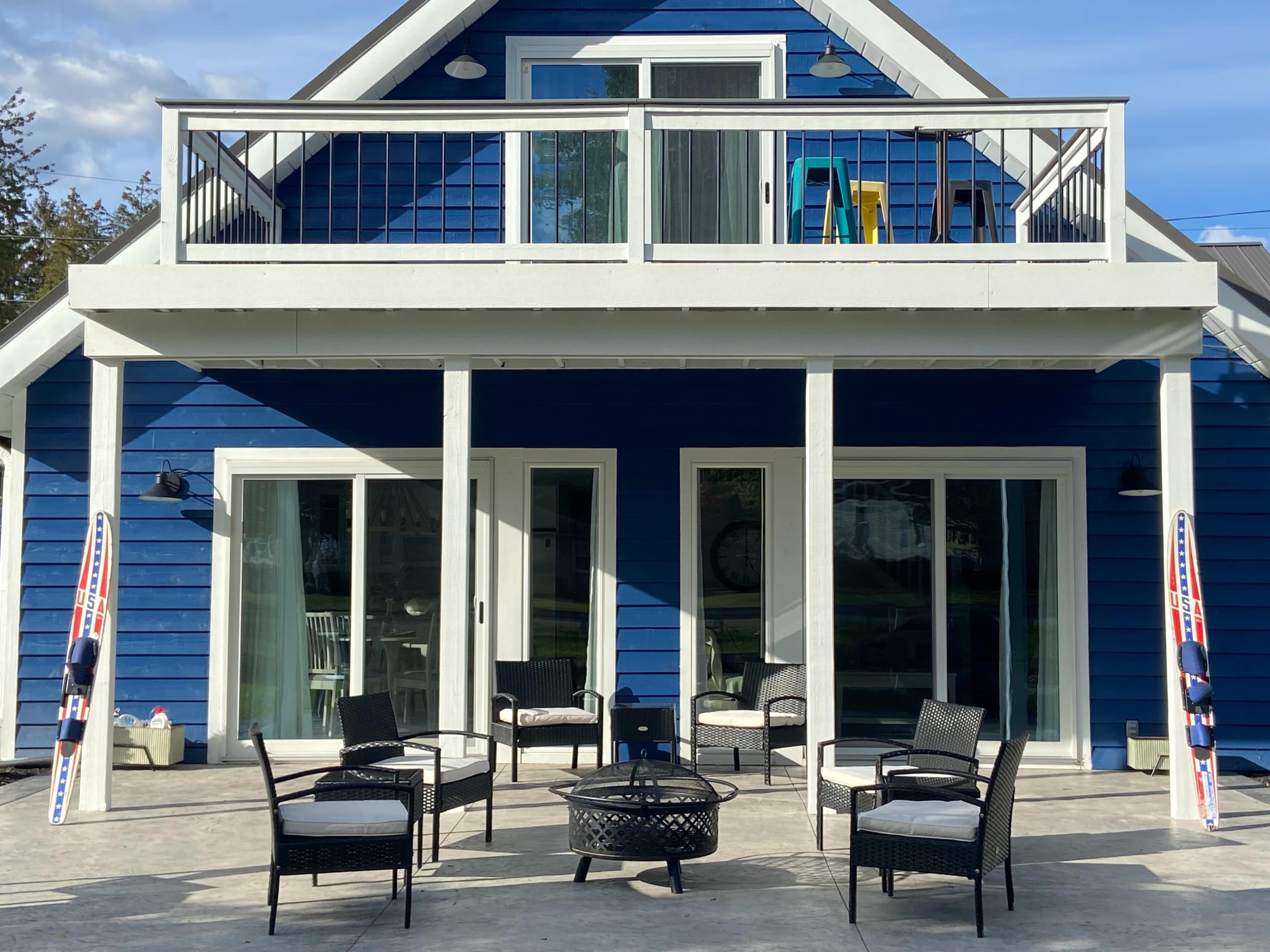 A blue two-story house with a balcony, surrounded by outdoor seating arrangements and a fire pit on a concrete patio.