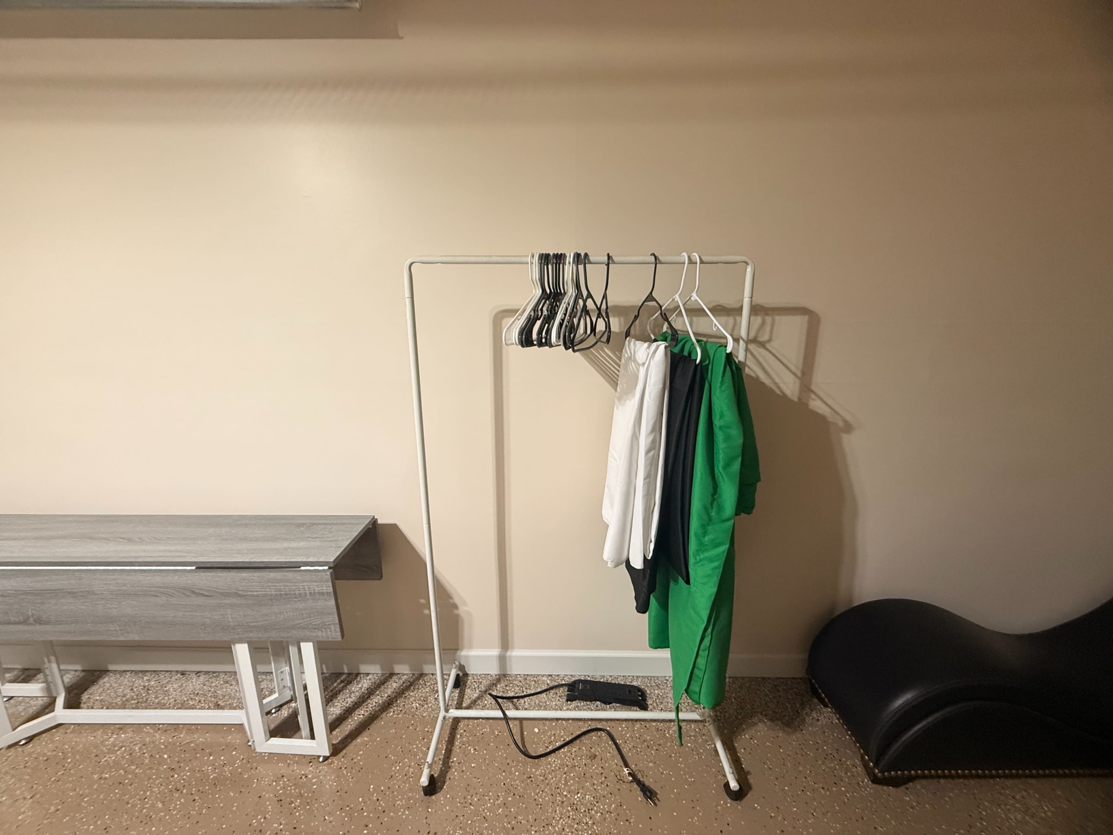 A clothing rack stands against a beige wall, holding several hangers with clothes, including a white shirt and a green garment, beside a gray bench and a black chair.