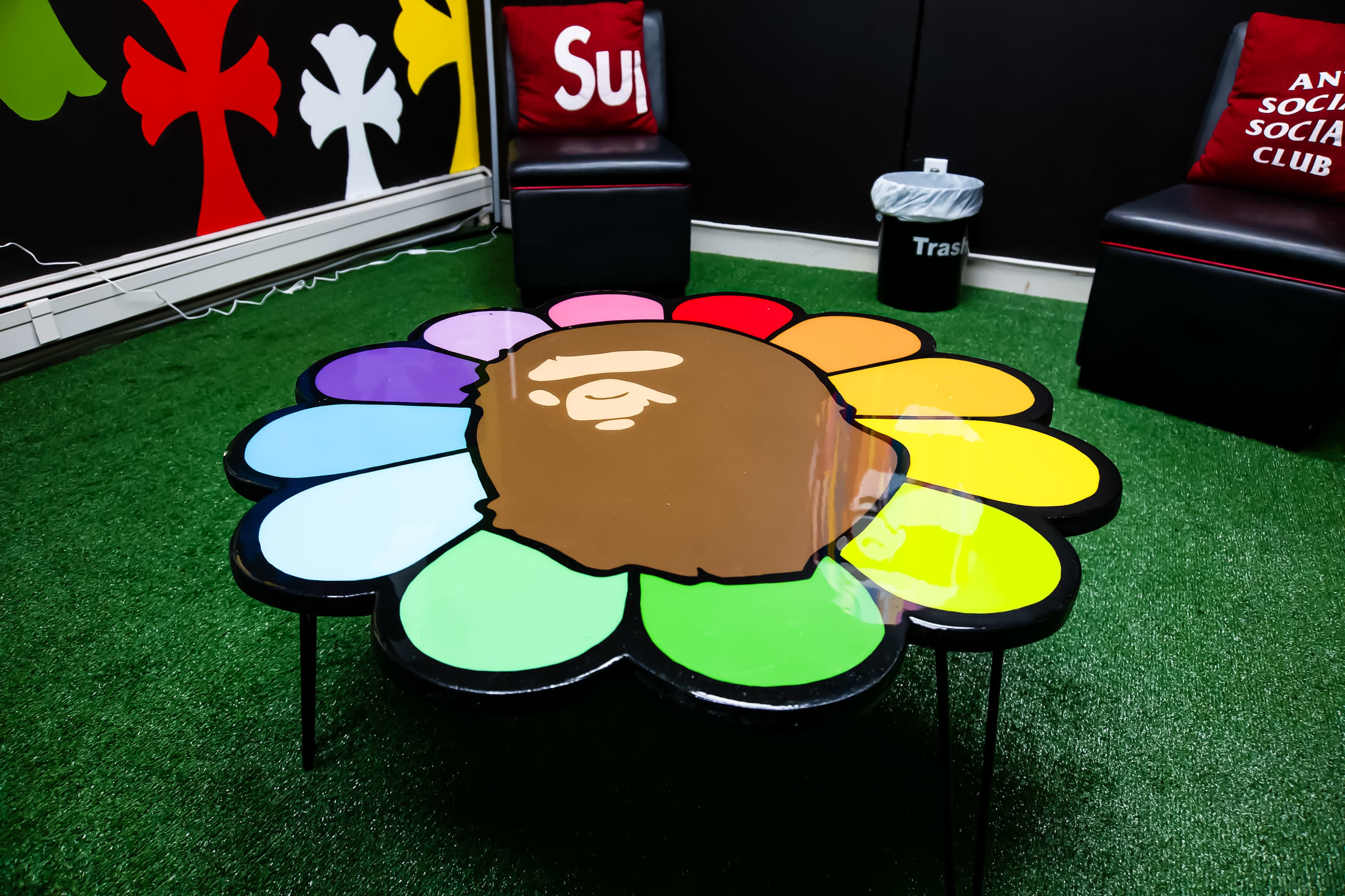 The image shows a colorful flower-shaped coffee table with a cartoon character's face in the center, set in a room with green turf flooring and two chairs with red throw pillows.