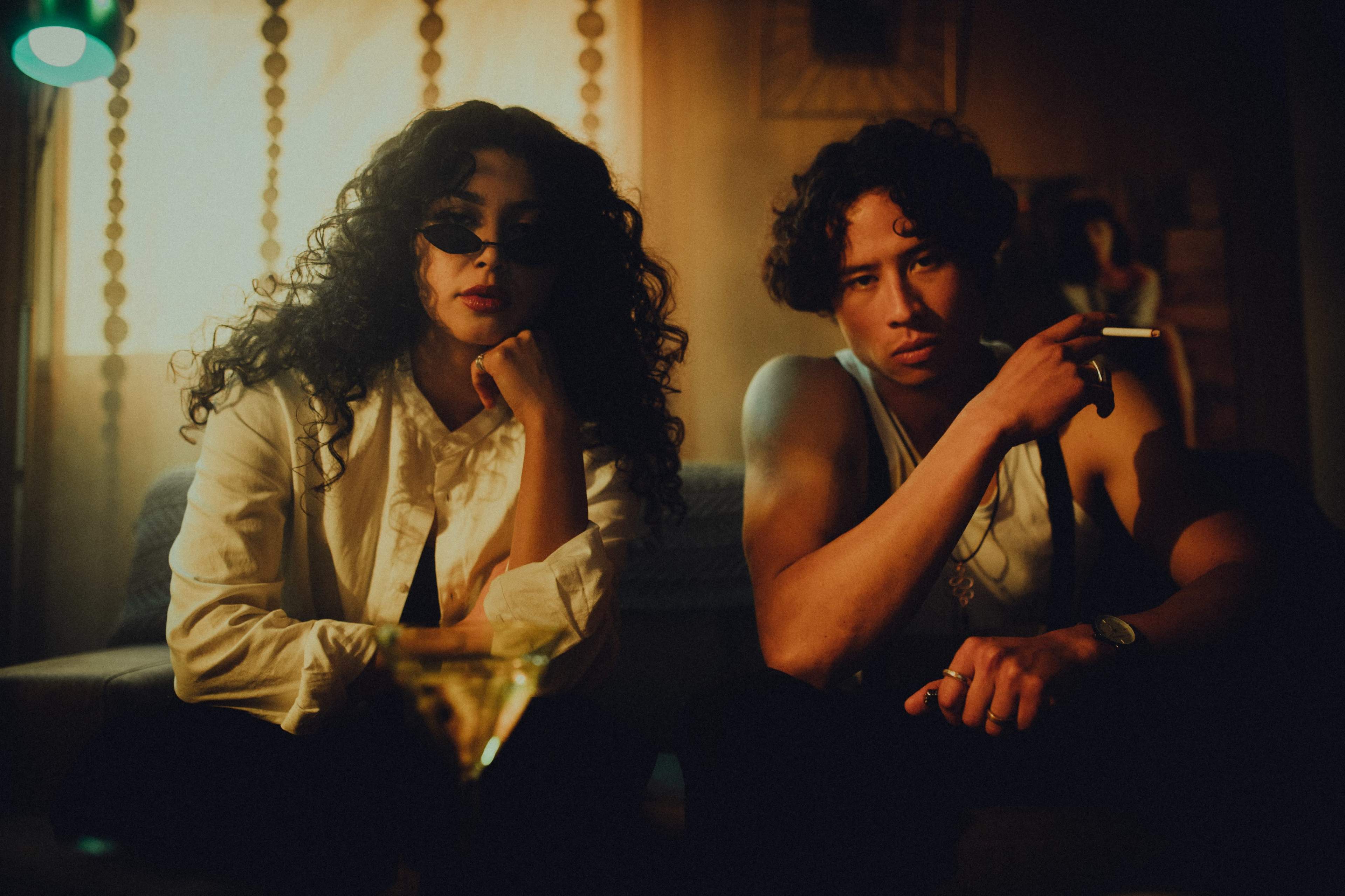 A man and a woman sit together on a couch in a dimly lit room, with the woman wearing sunglasses and the man holding a cigarette.