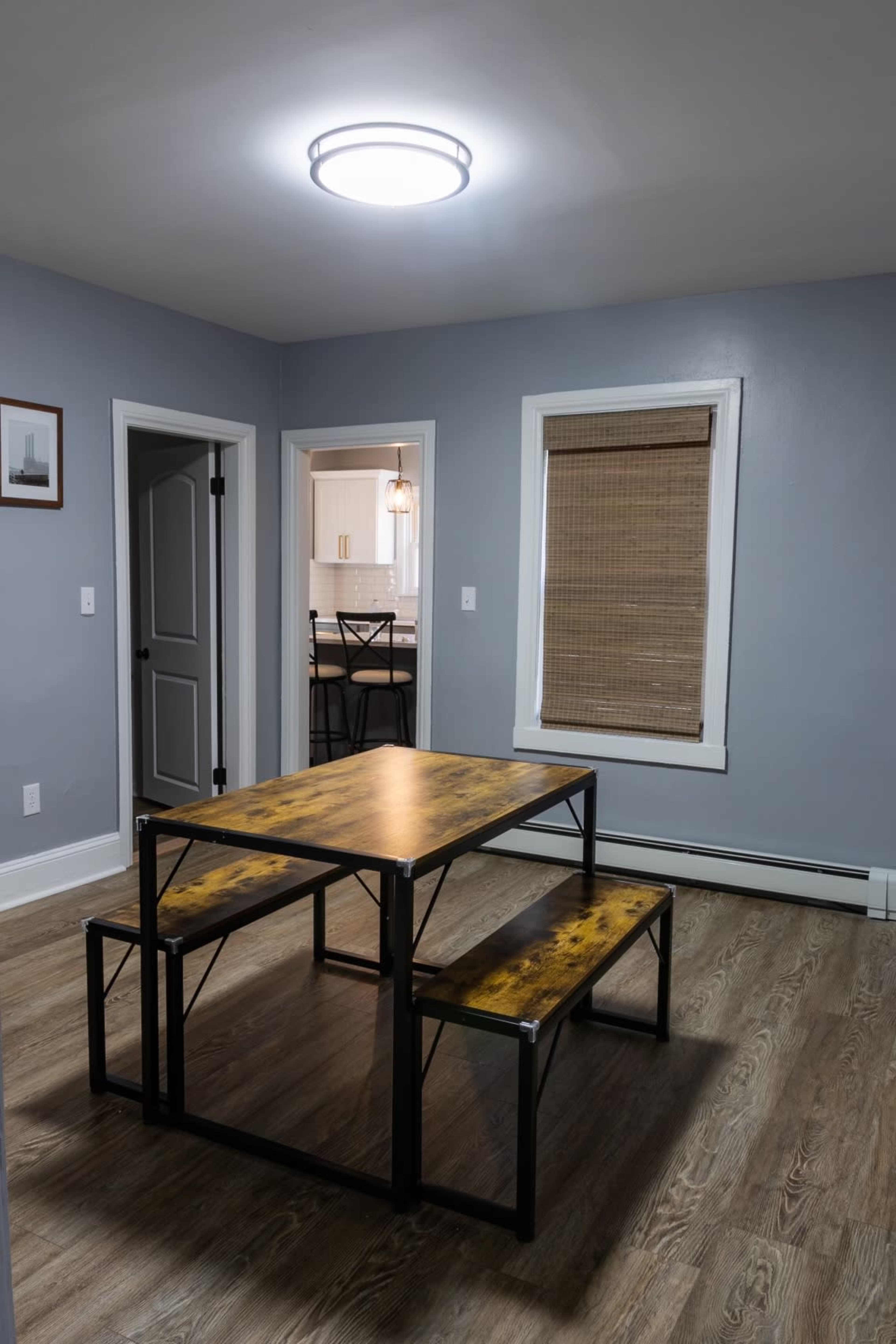 A wooden table with metal benches sits in a simple dining area, illuminated by a round ceiling light, with a doorway leading to a kitchen in the background.